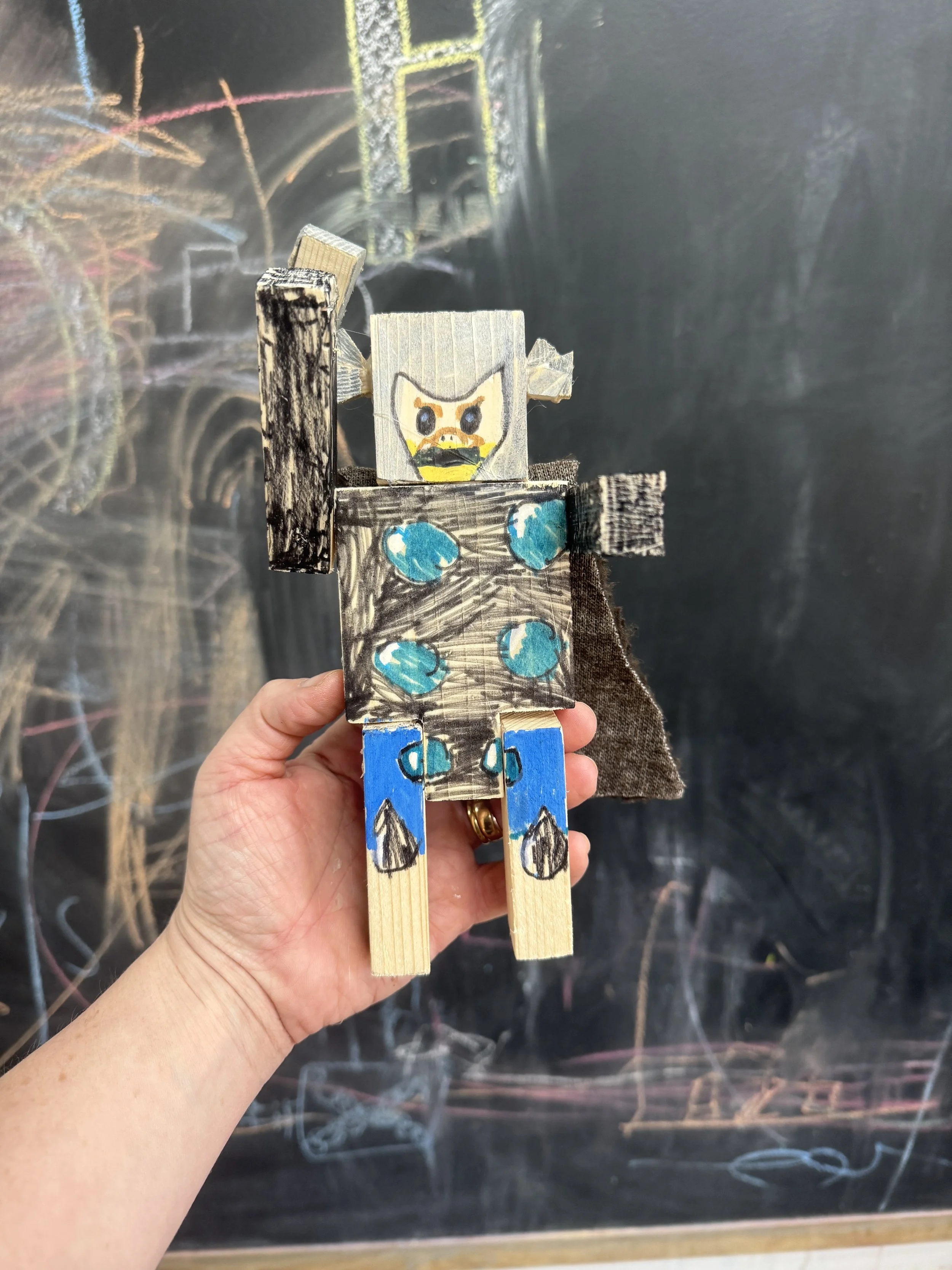 Hand holding a paper craft figure resembling a cartoon character with spiky hair, a dark beard, and wearing a blue and black outfit, in front of a blackboard with colorful chalk drawings.