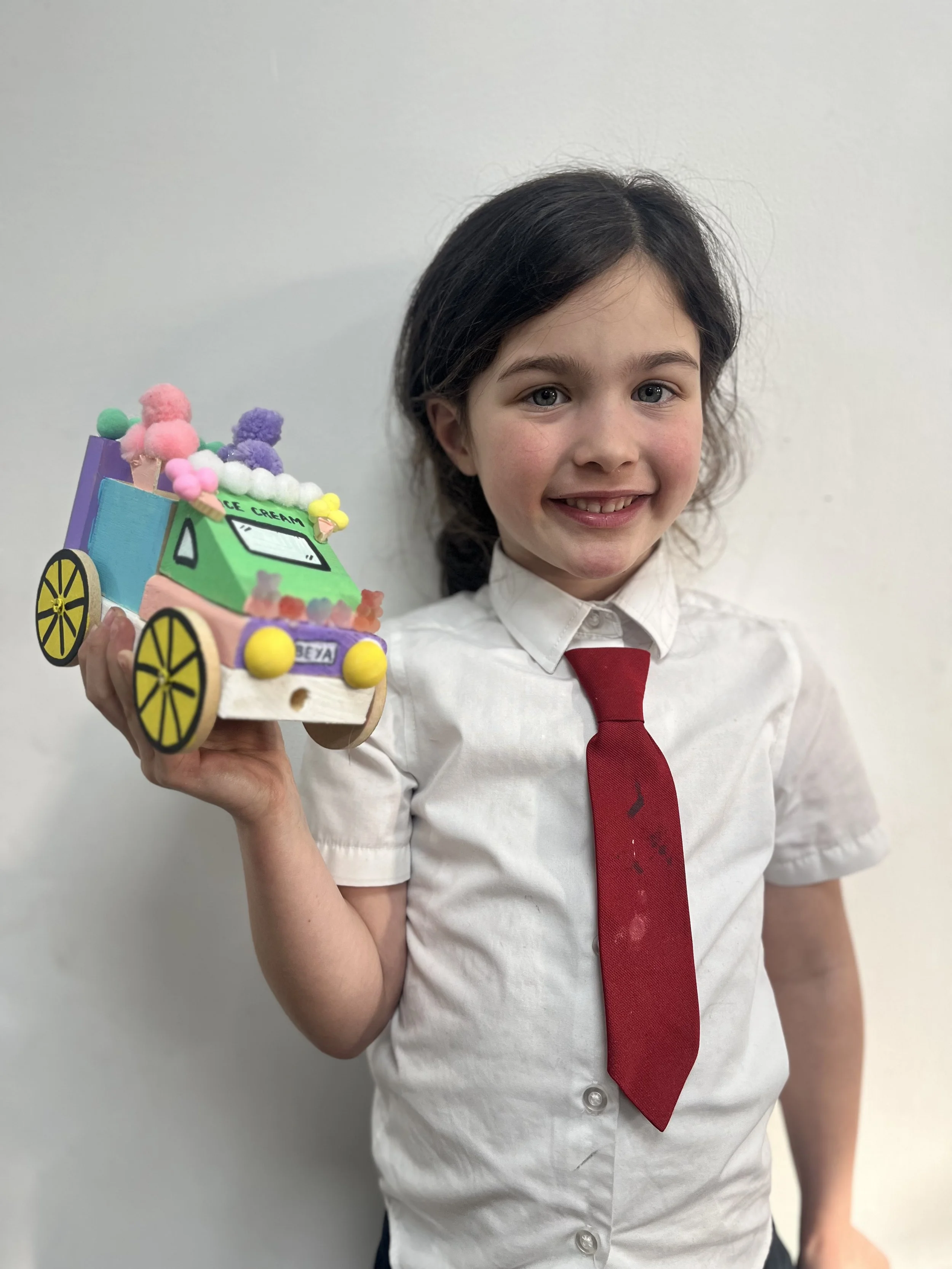 A young girl with dark hair, smiling, wearing a white shirt and a red tie, holding a colorful, handcrafted ice cream truck made of craft materials.