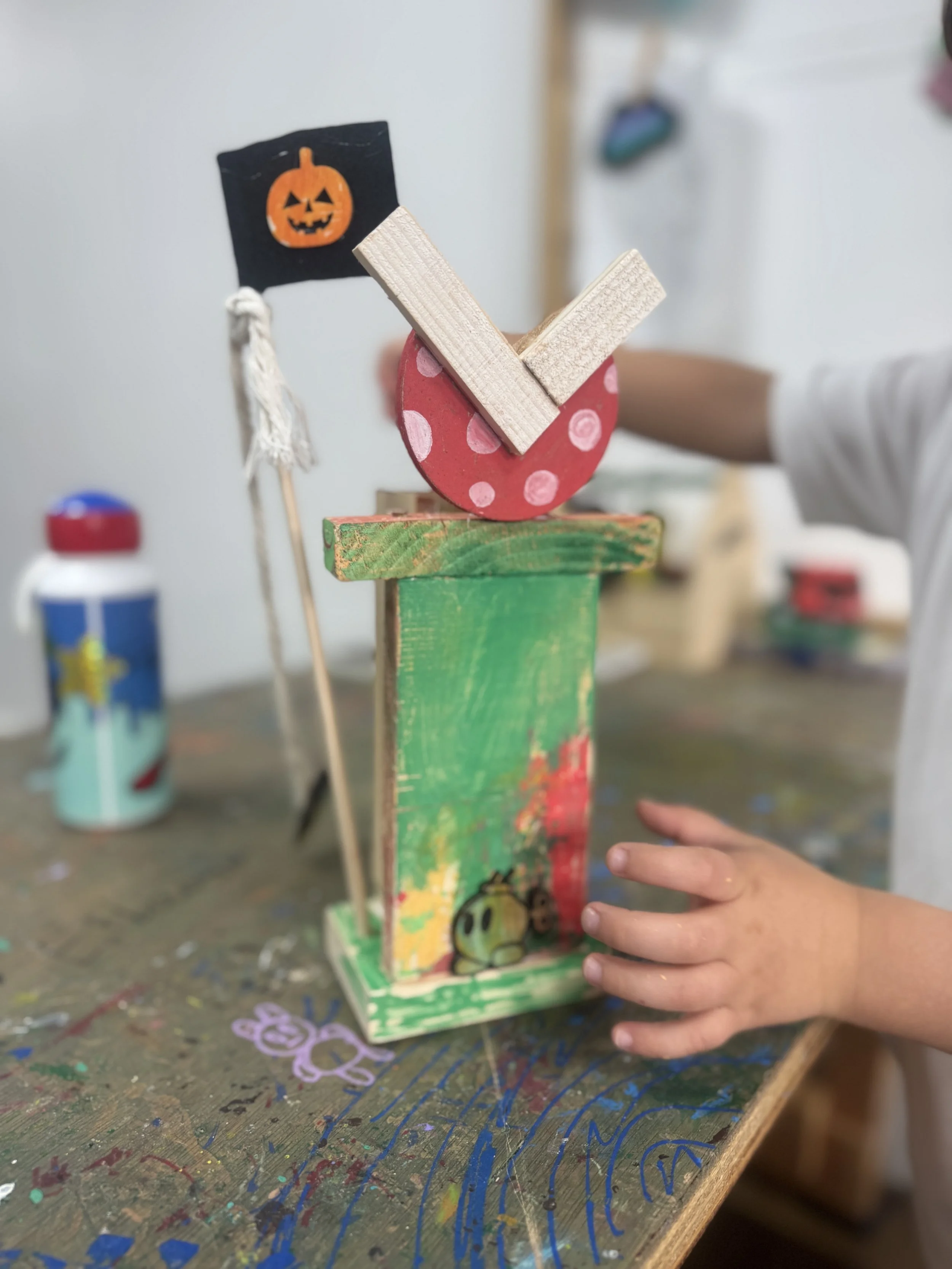 Child's colorful craft project resembling a pumpkin with a flag that has a jack-o'-lantern face, on a cluttered wooden table with paint and drawings.