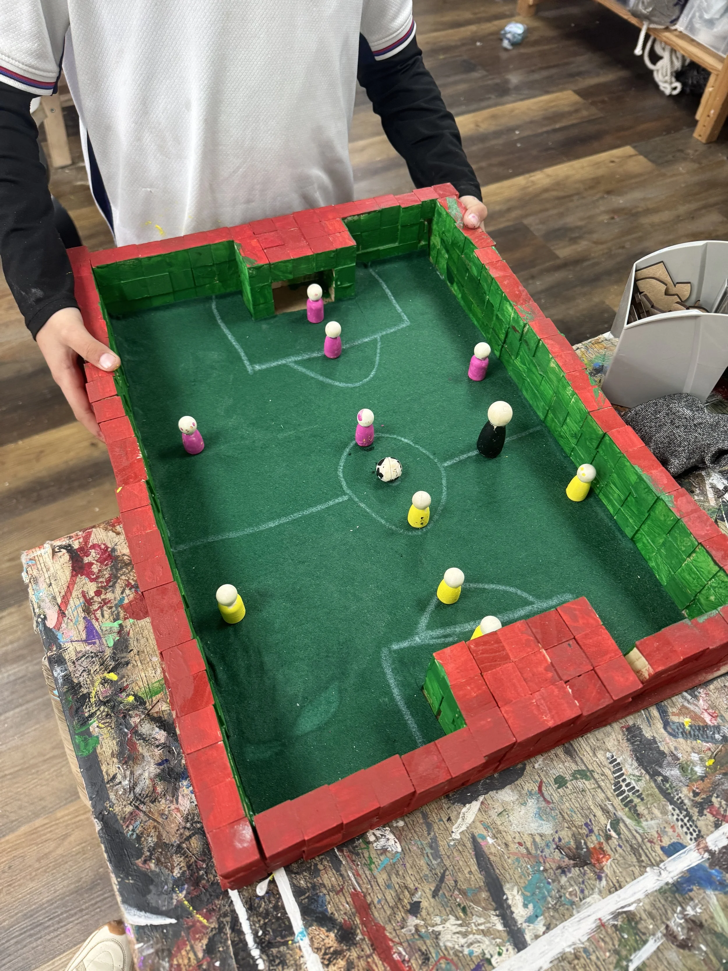 A handmade mini soccer game with a green felt field, red and green painted cardboard walls, small figurines as players, and a small soccer ball as the ball, set on a paint-stained table.