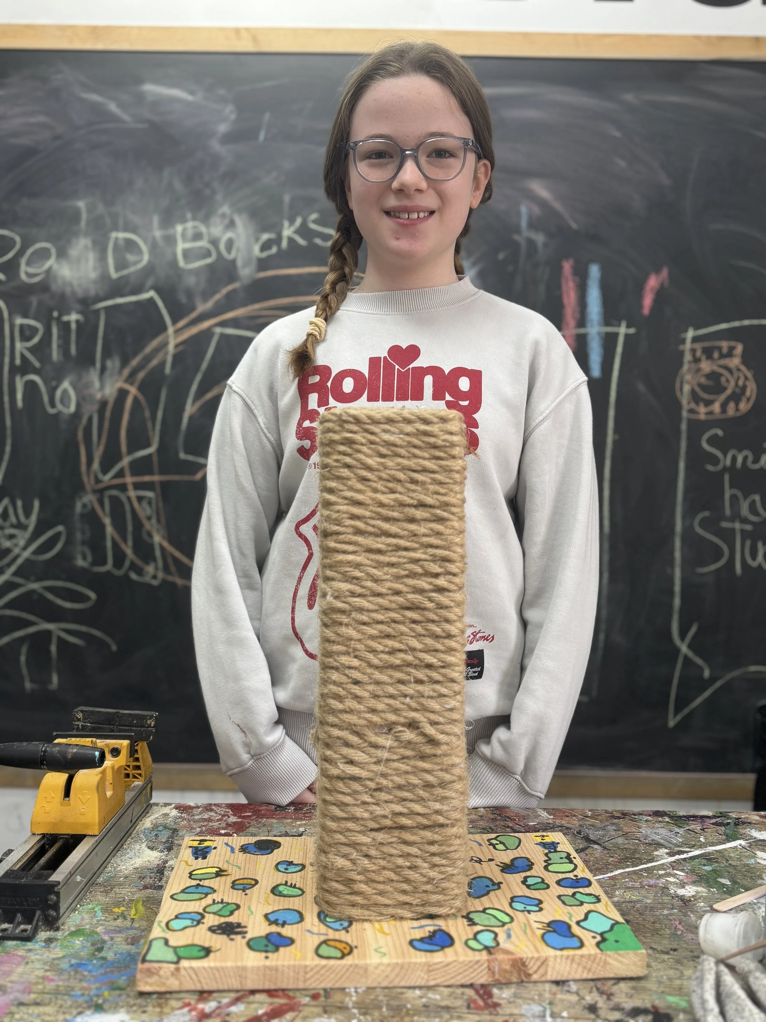 Girl with glasses standing behind a tall, rectangular piece of rope art on a painted wooden board in a classroom with a blackboard and drawings in the background.