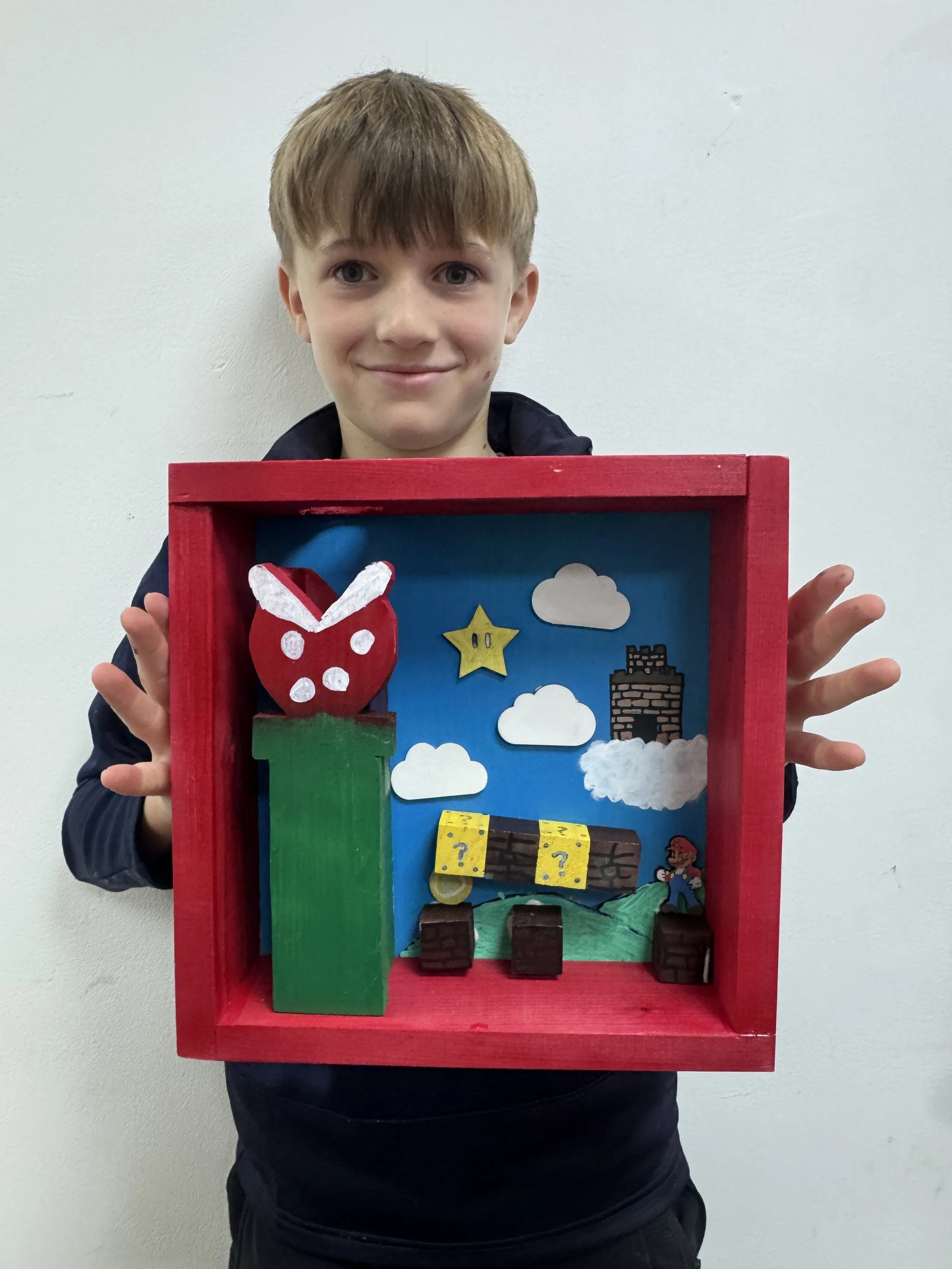 A young boy holding a red shadow box display with Super Mario-themed decorations including a green pipe, a red apple with white eyes and ears, clouds, a star, a brick block, a question block, and a small Mario figure.