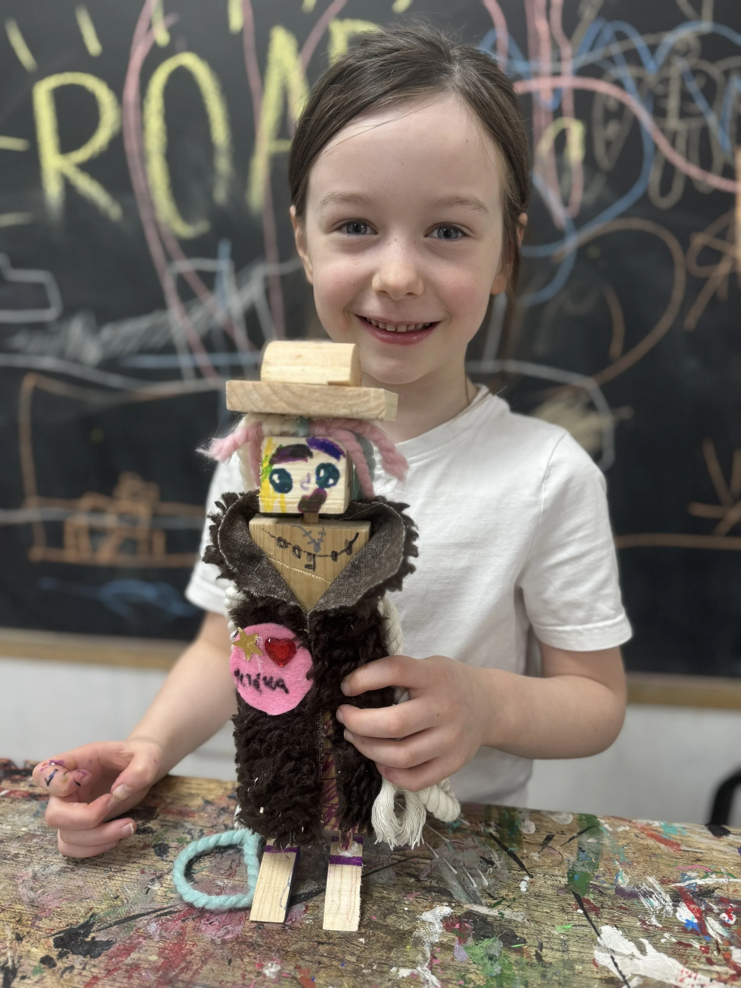 A young girl smiling and holding a handmade doll in a classroom with a blackboard in the background.