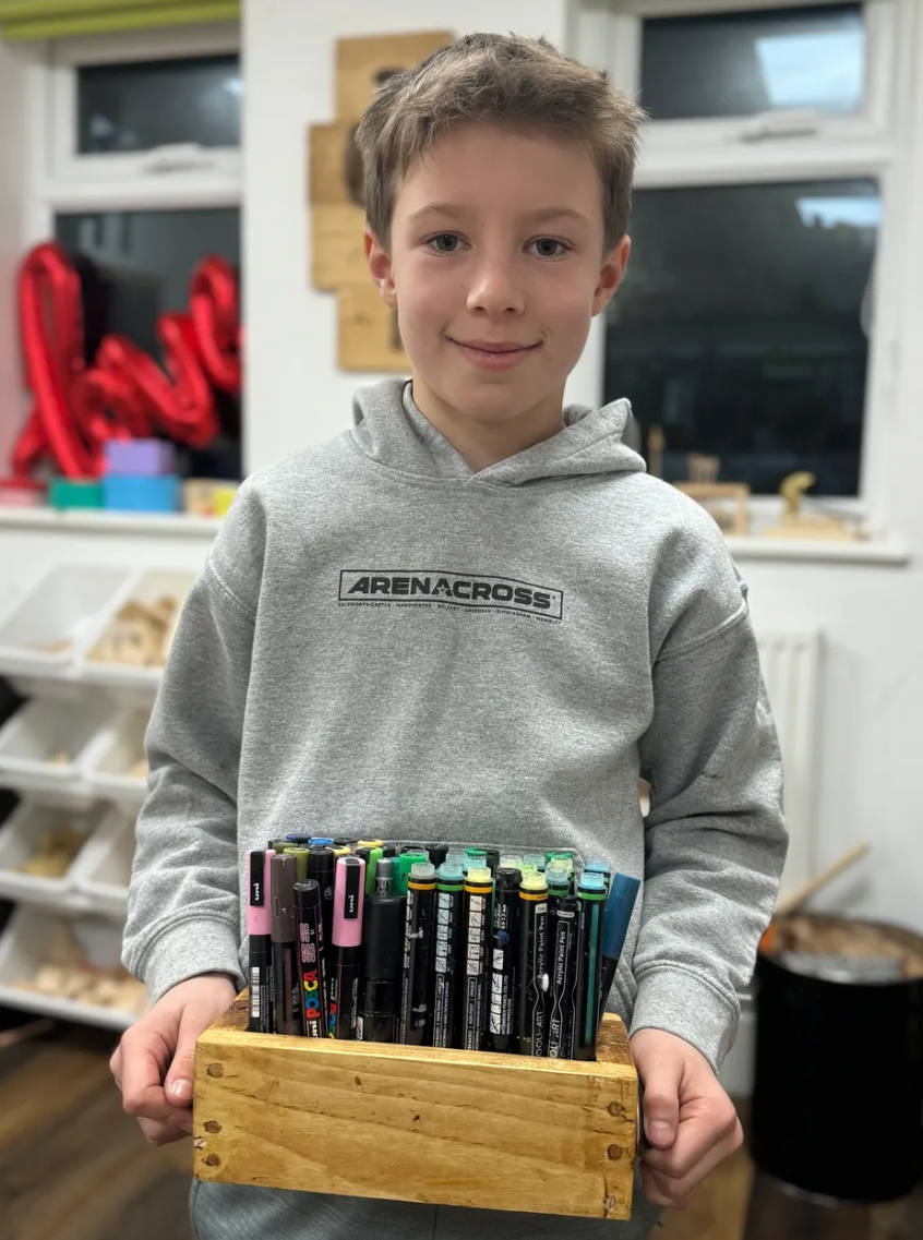 A young boy with light brown hair wearing a gray hoodie that says 'ARENA CROSS' is holding a wooden box filled with various colored pens and markers. The background shows a room with white walls, a window, and some shelves with storage bins and decor