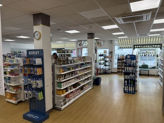 Interior of a pharmacy or convenience store with shelves stocked with various health and personal care products, a clock on a pillar, and a large window in the background.