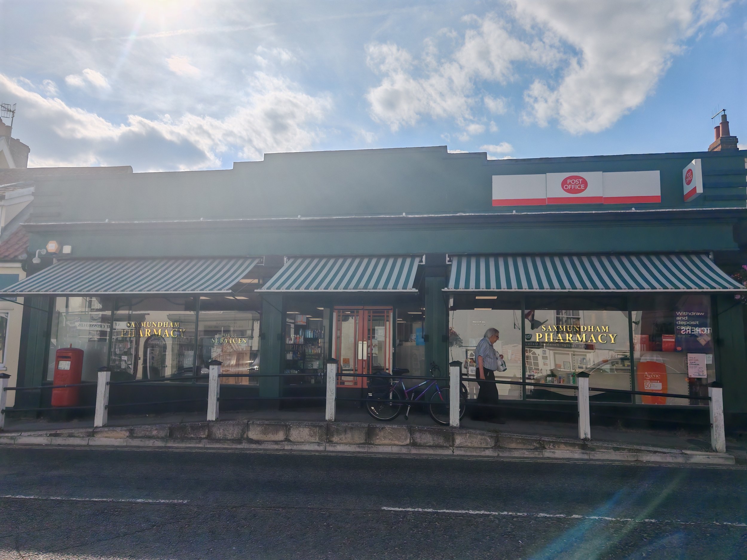A storefront with a sign that reads 'Saxmundham Pharmacy' and an awning with blue and white stripes. There is a person standing outside, and a bicycle is leaning against a white fence.