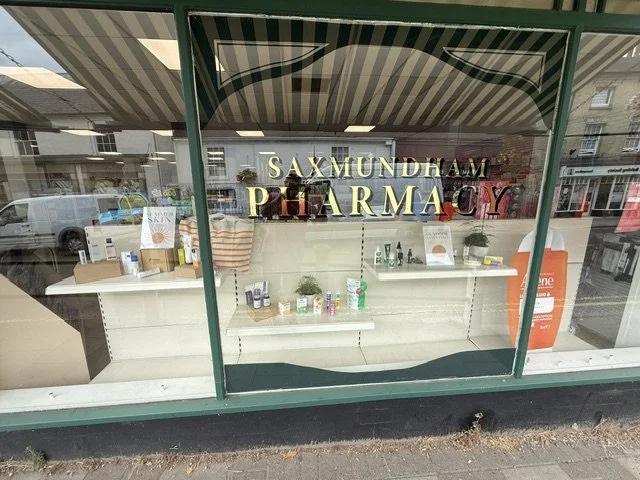 Storefront window display with the sign 'Saxmundham Pharmacy' in gold letters, showing shelves with medications and health products inside.