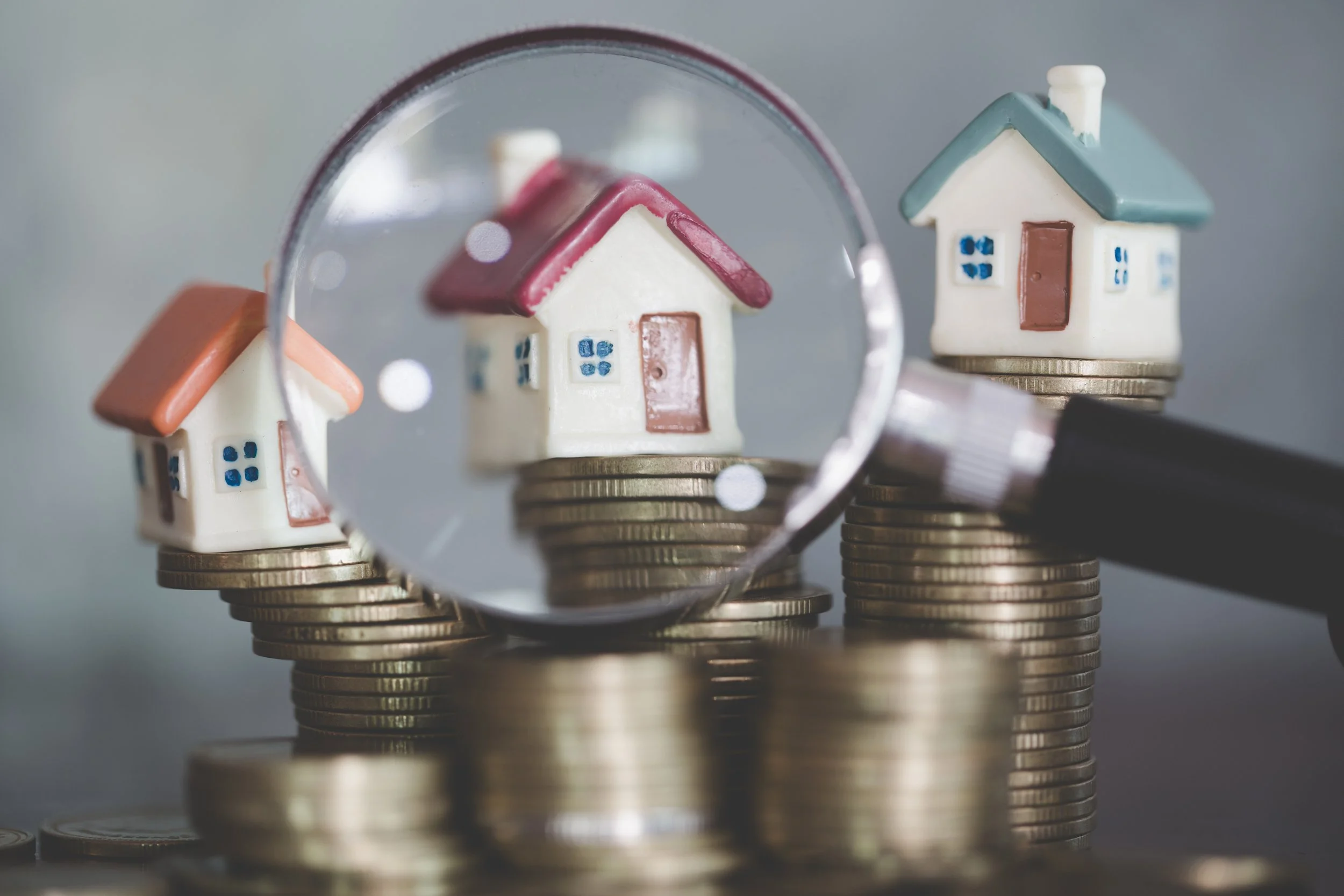 Miniature houses on stacked coins viewed through a magnifying glass, symbolizing real estate investment or housing market analysis.