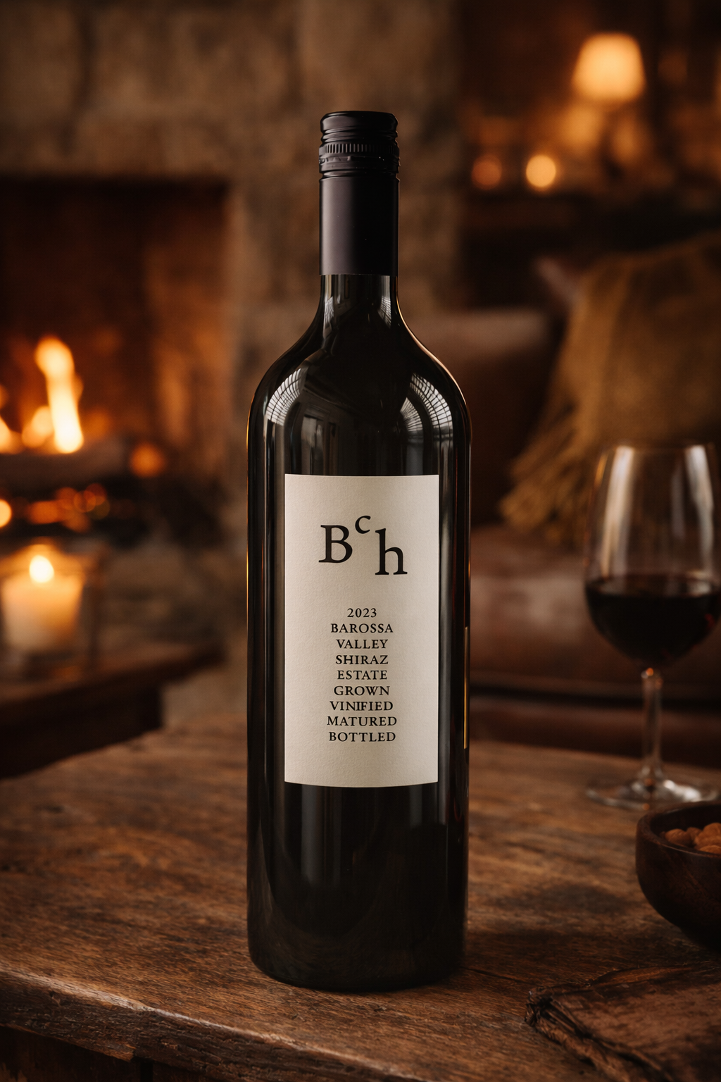 A bottle of 2023 Barossa Valley Shiraz wine on a rustic wooden table inside a cozy room with a stone fireplace and warm lighting, with a glass of red wine and a bowl of snacks nearby.