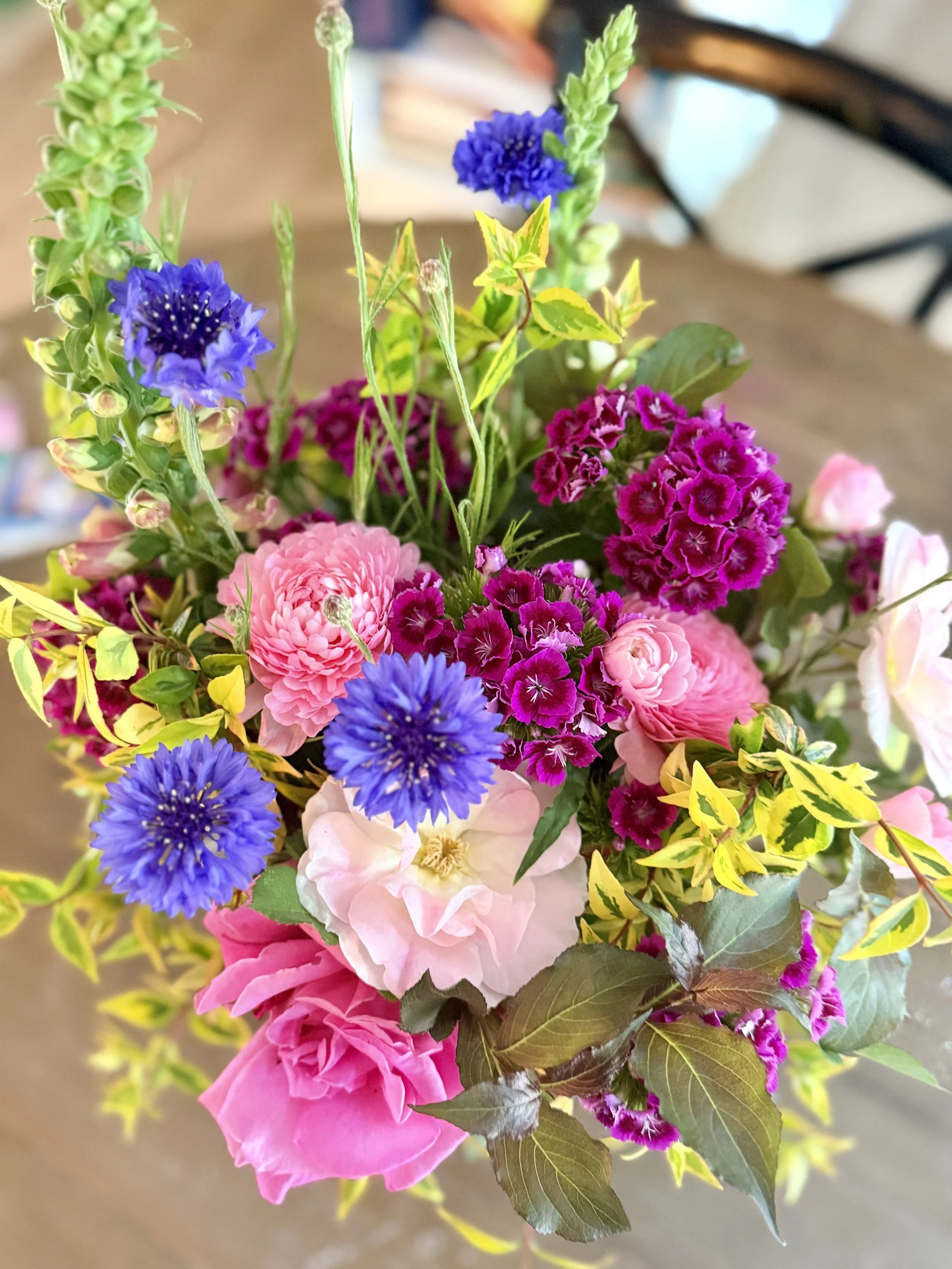 A colorful bouquet of flowers including pink roses, purple carnations, blue cornflowers, and yellow flowers with green leaves on a wooden surface.