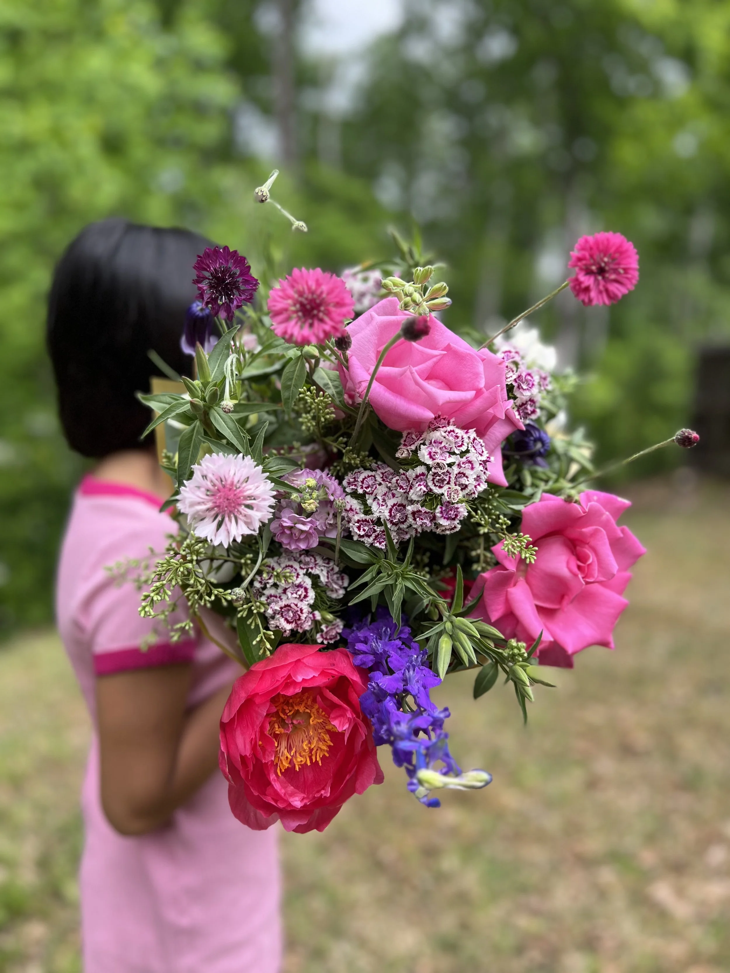 Child holding a colorful bouquet of pink, purple, and orange flowers in a blurred outdoor background.