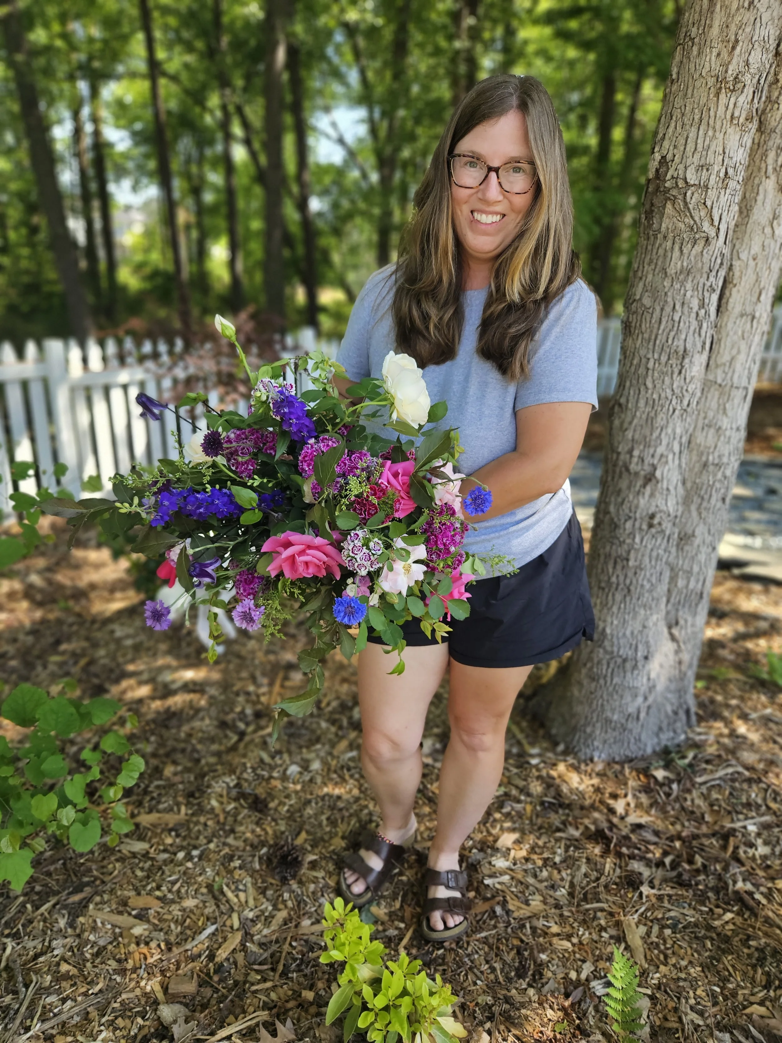 A woman with long brown hair, glasses, and a blue t-shirt stands outdoors near a tree, holding a large bouquet of colorful flowers, in a wooded area with sunlight filtering through the trees.