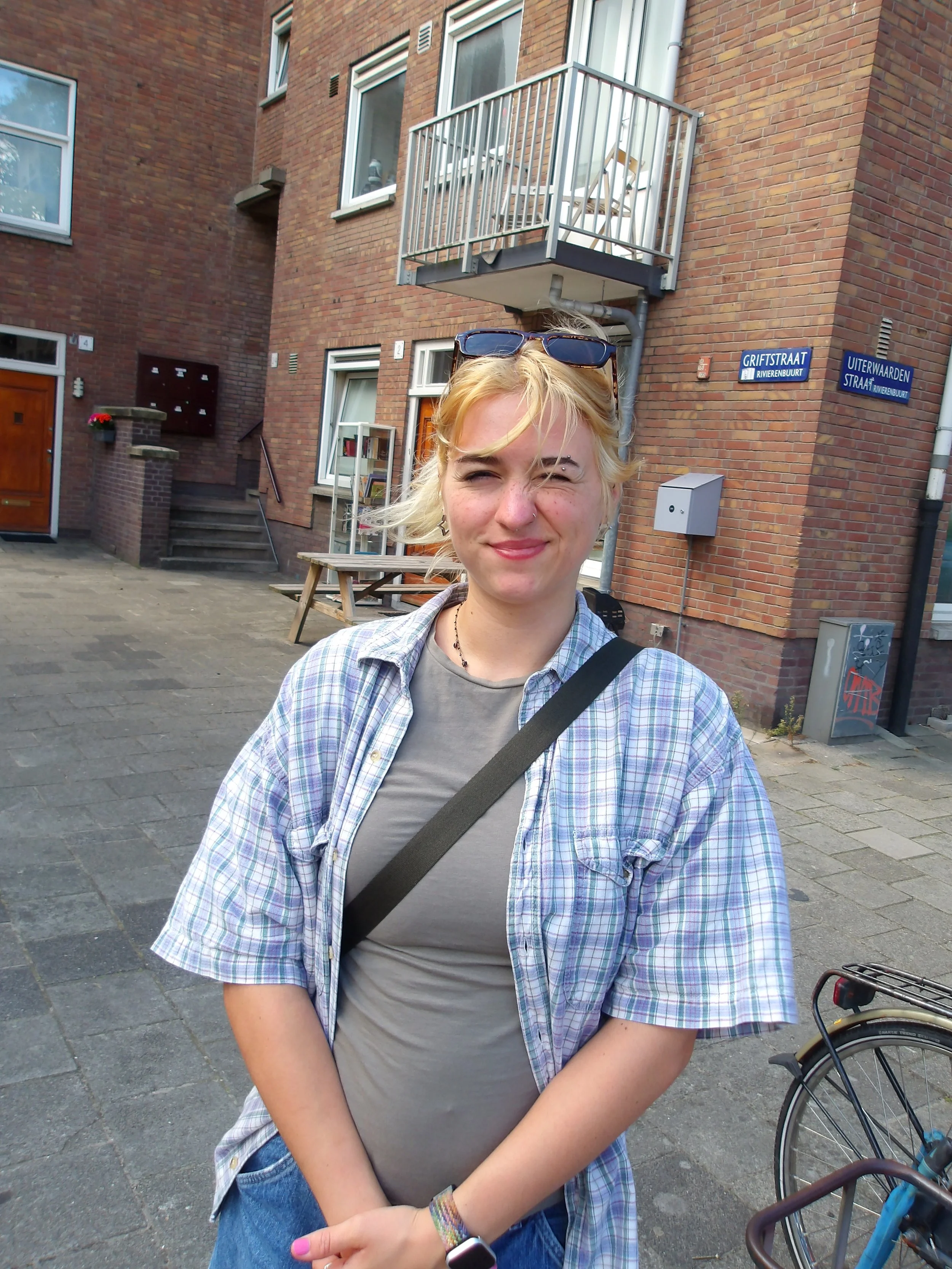 A young woman with blonde hair, sunglasses on her head, and a nose piercing smiling outdoors in front of a brick building with a balcony, street signs, and a bicycle.