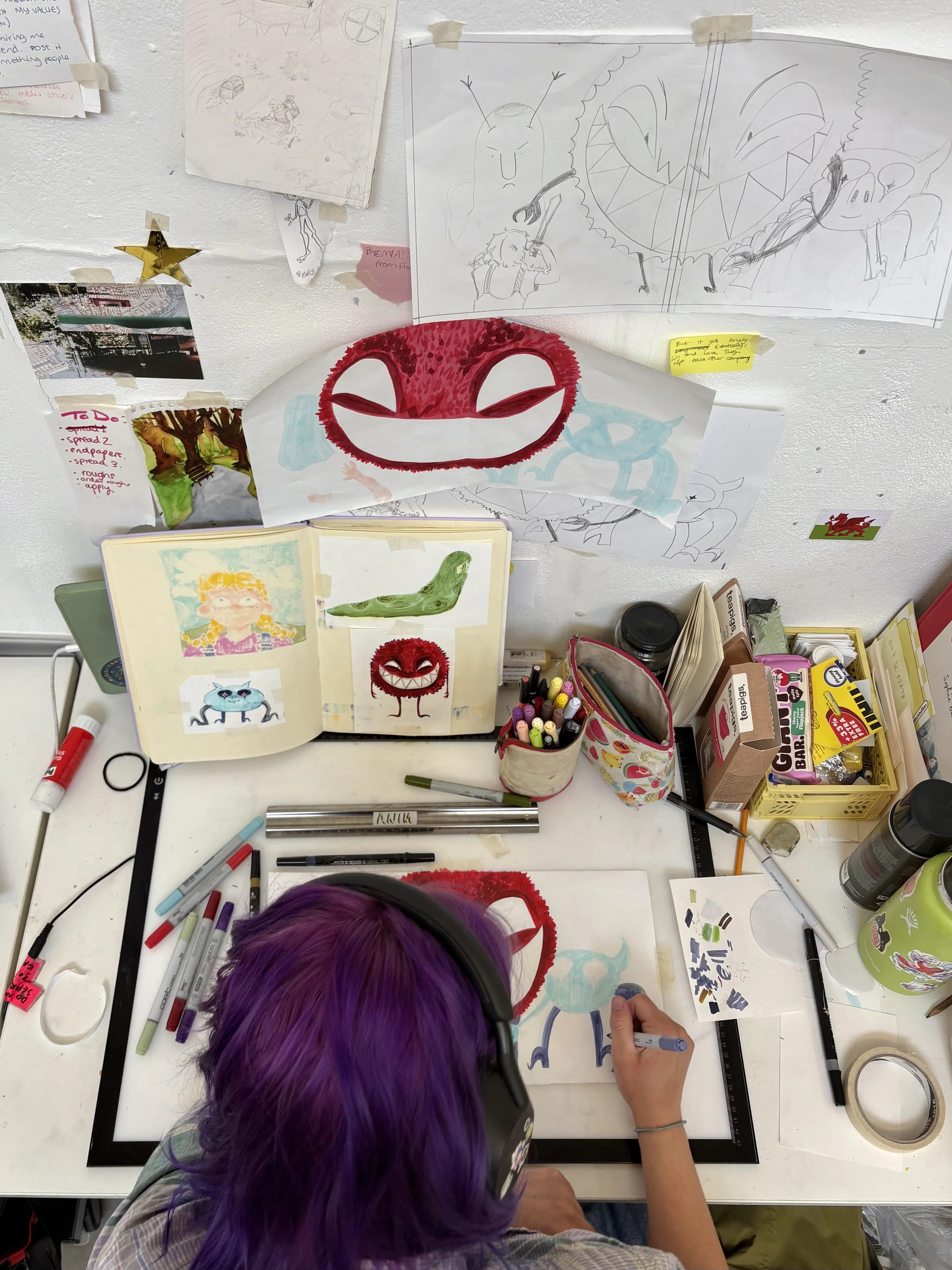 A person with purple hair wearing headphones drawing sketches of cartoon characters and creatures on paper, surrounded by markers, pens, and various art supplies on a cluttered white desk.