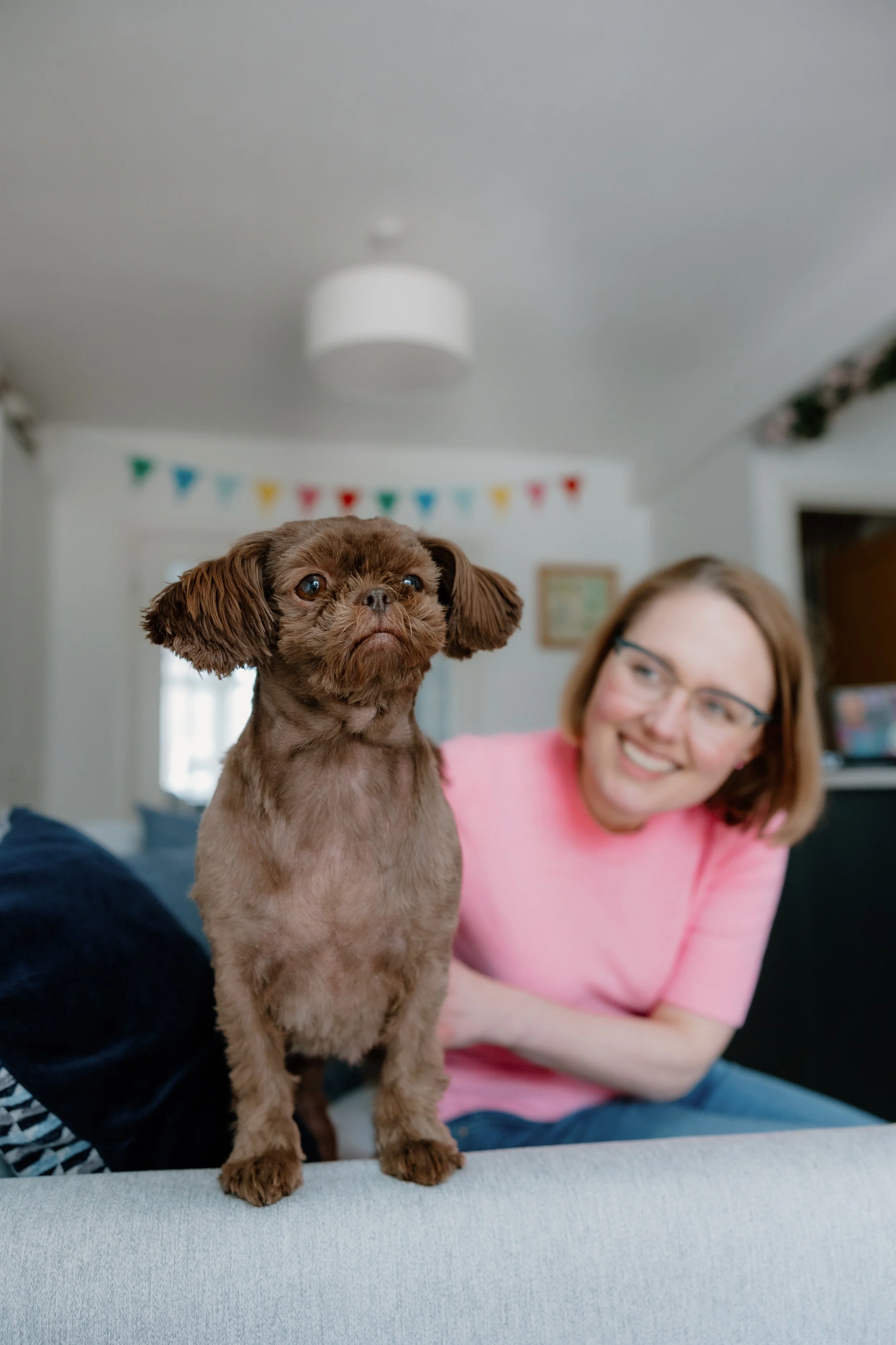 a little furry brown dog int he forefront of the image, and behind it, a woman in glasses smiling at the dog