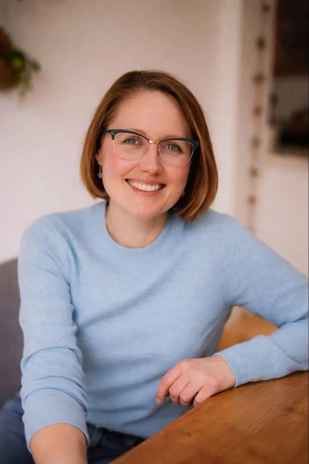Smiling woman with a blonde bob haircut and glasses, wearing a light blue sweater and leaning on a wooden table.