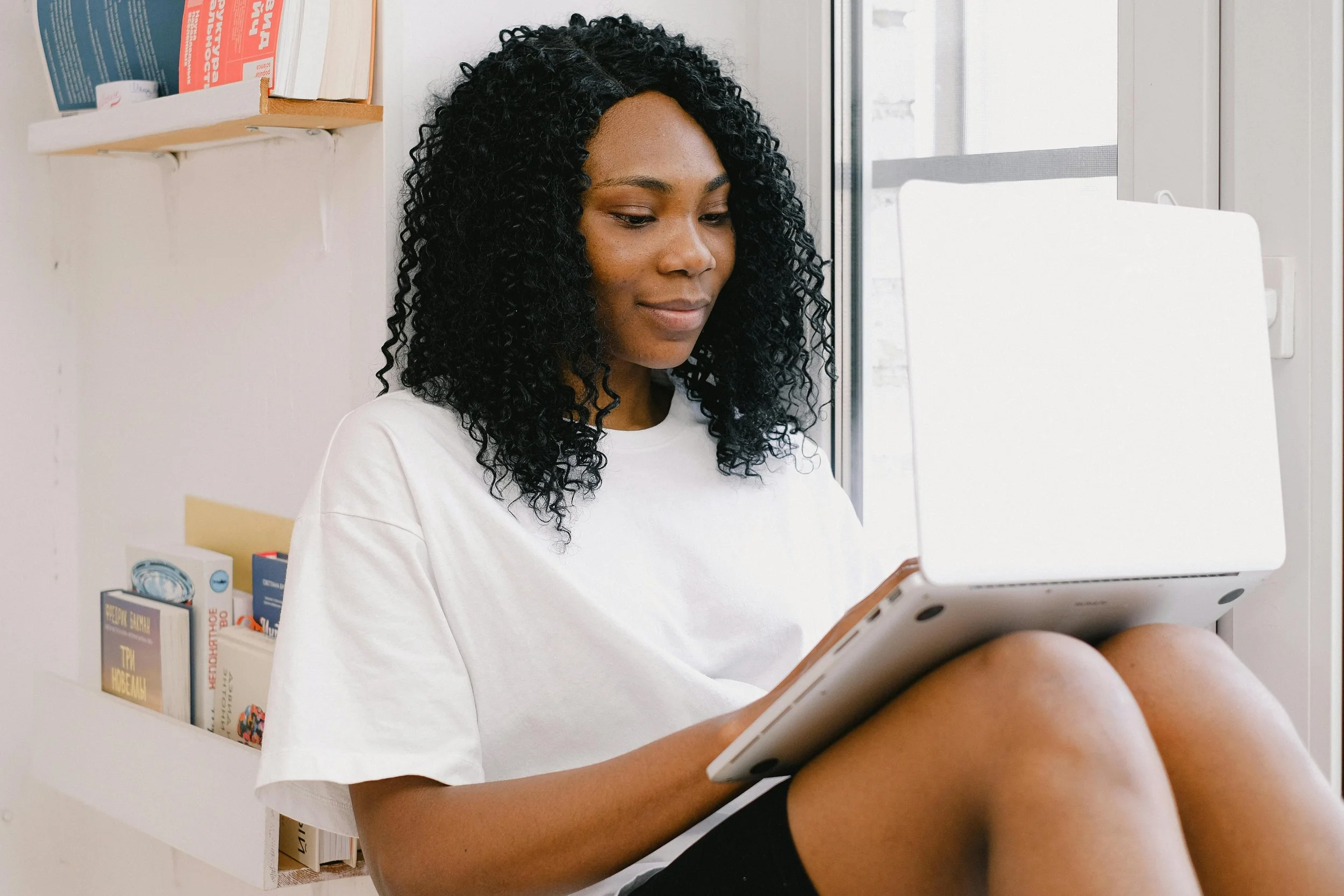 Woman with black curly hair sitting on a chair, balancing a laptop on her lap