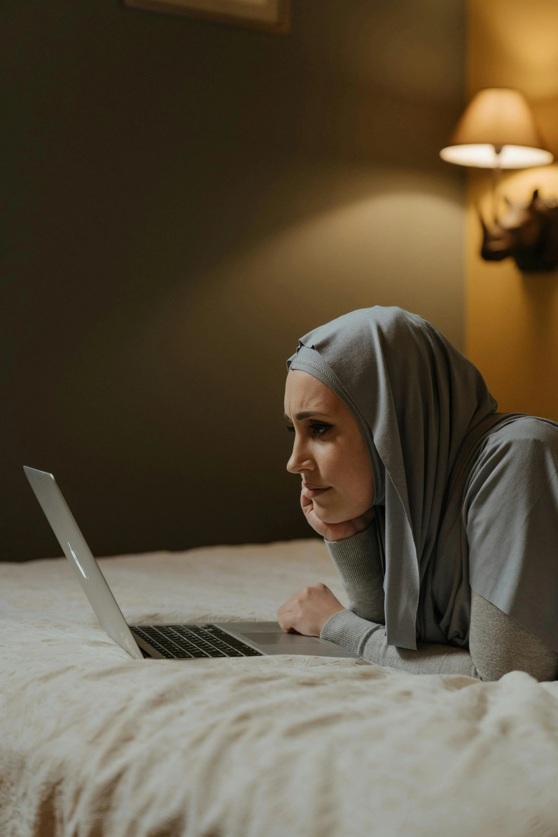 woman in a headscarf lying on her stomach on a bed, looking at a laptop screen