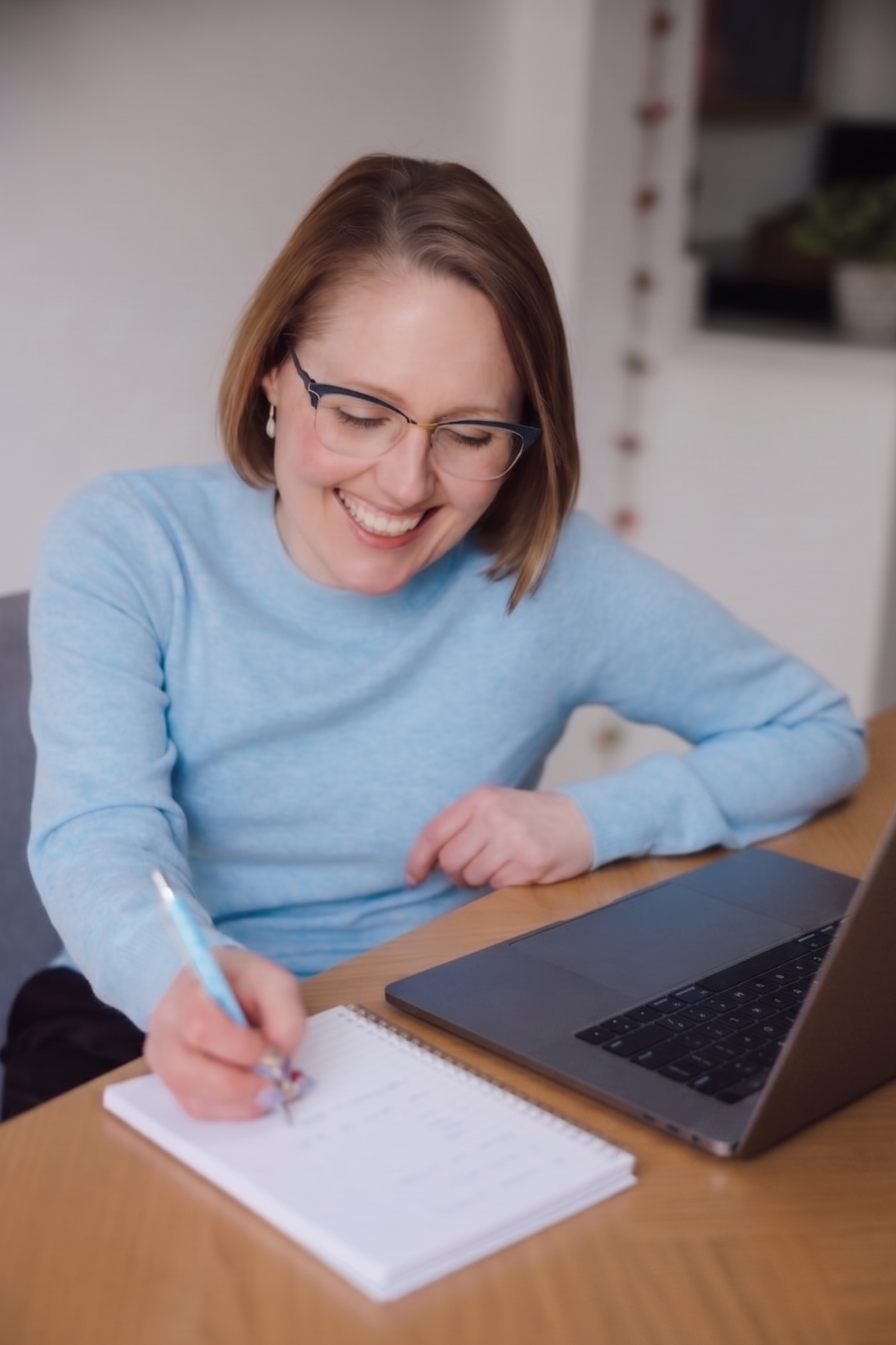 Woman in blue jumper, sitting at table writing with a laptop in front of her