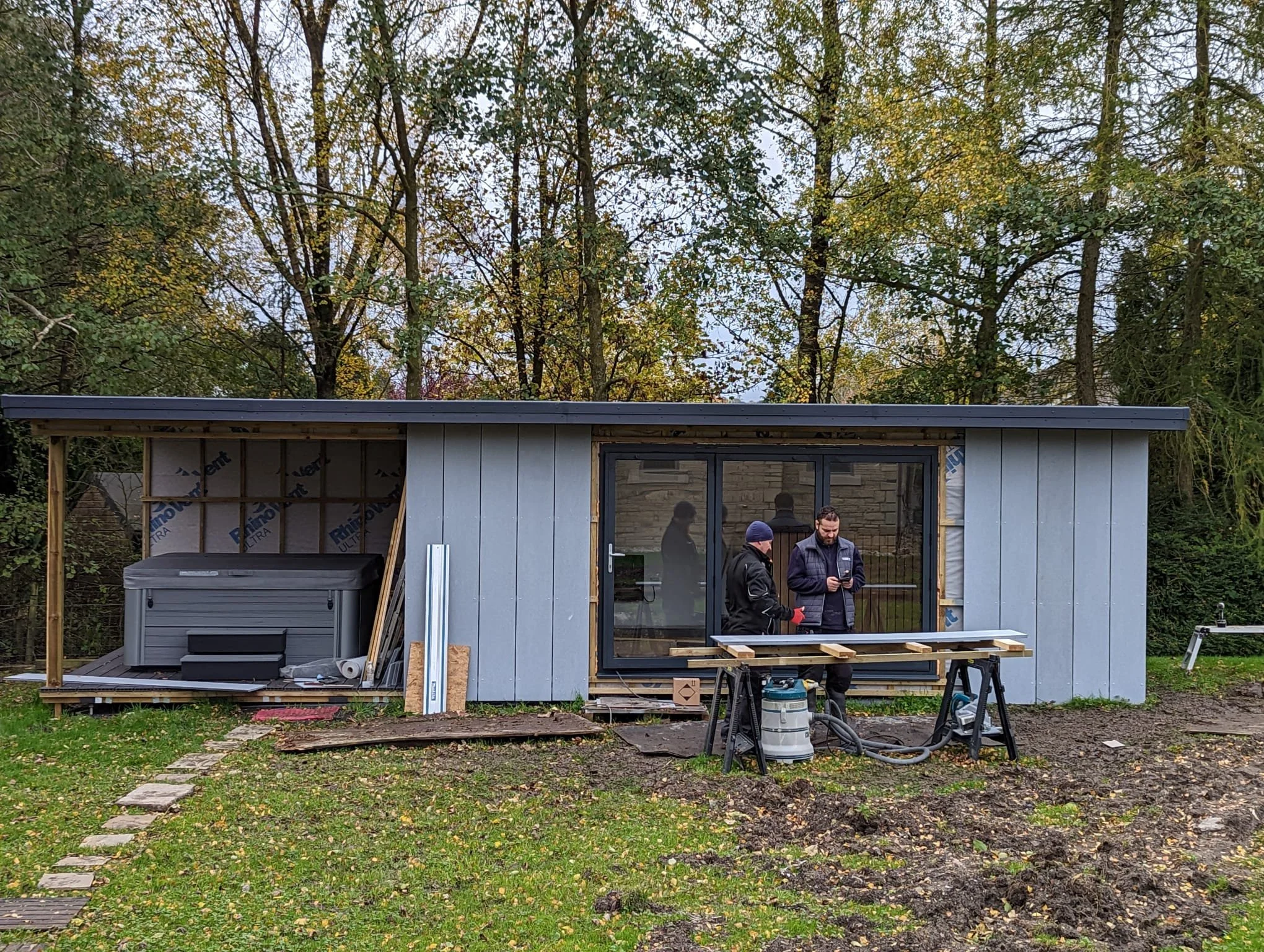 Two men in hoodies and jackets working on a small house under construction in a backyard, with trees in the background and construction tools and materials around.