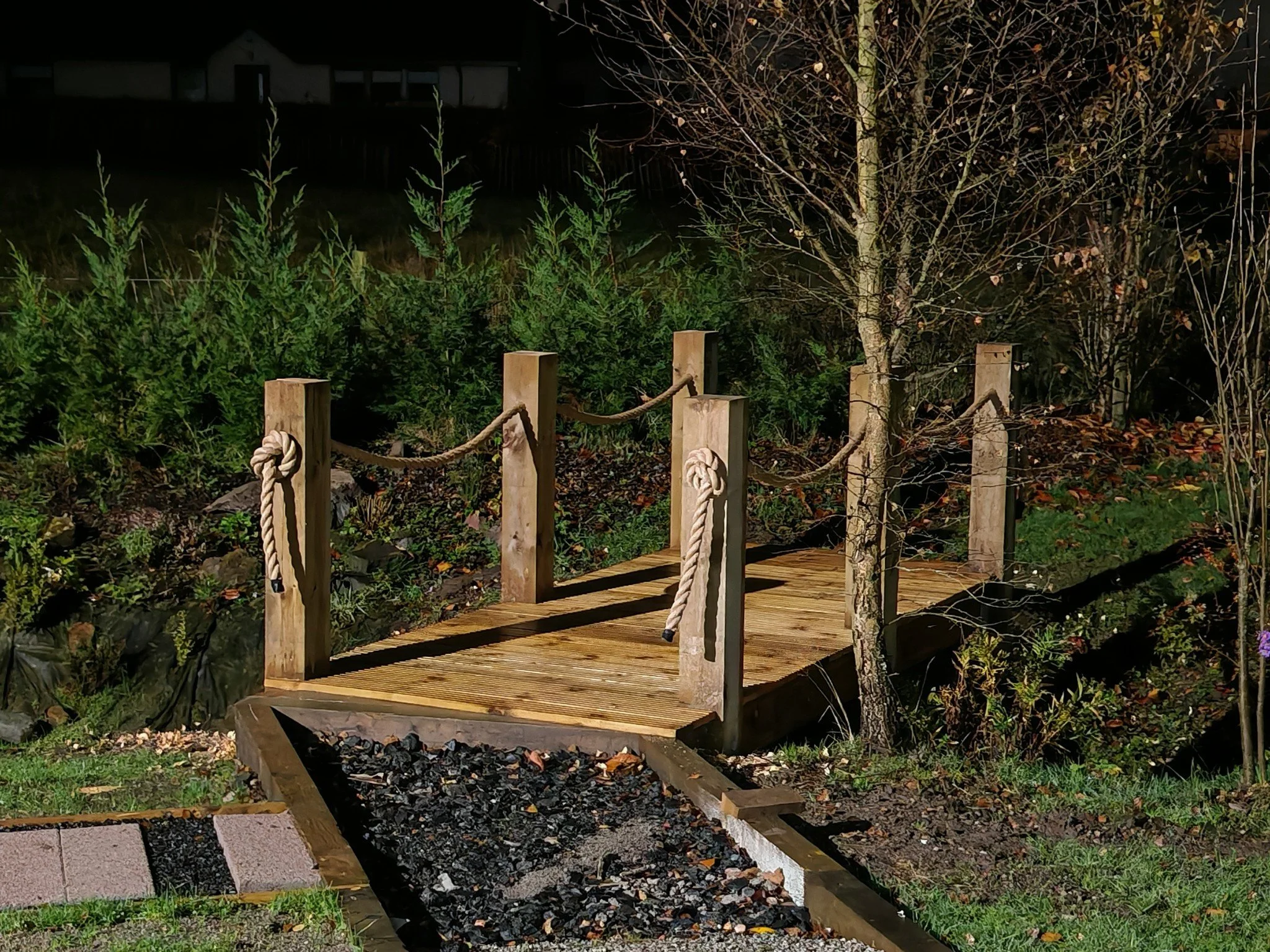 Nighttime view of a small wooden deck with a natural rope railing, surrounded by evergreen and deciduous trees, with a dark sky in the background.