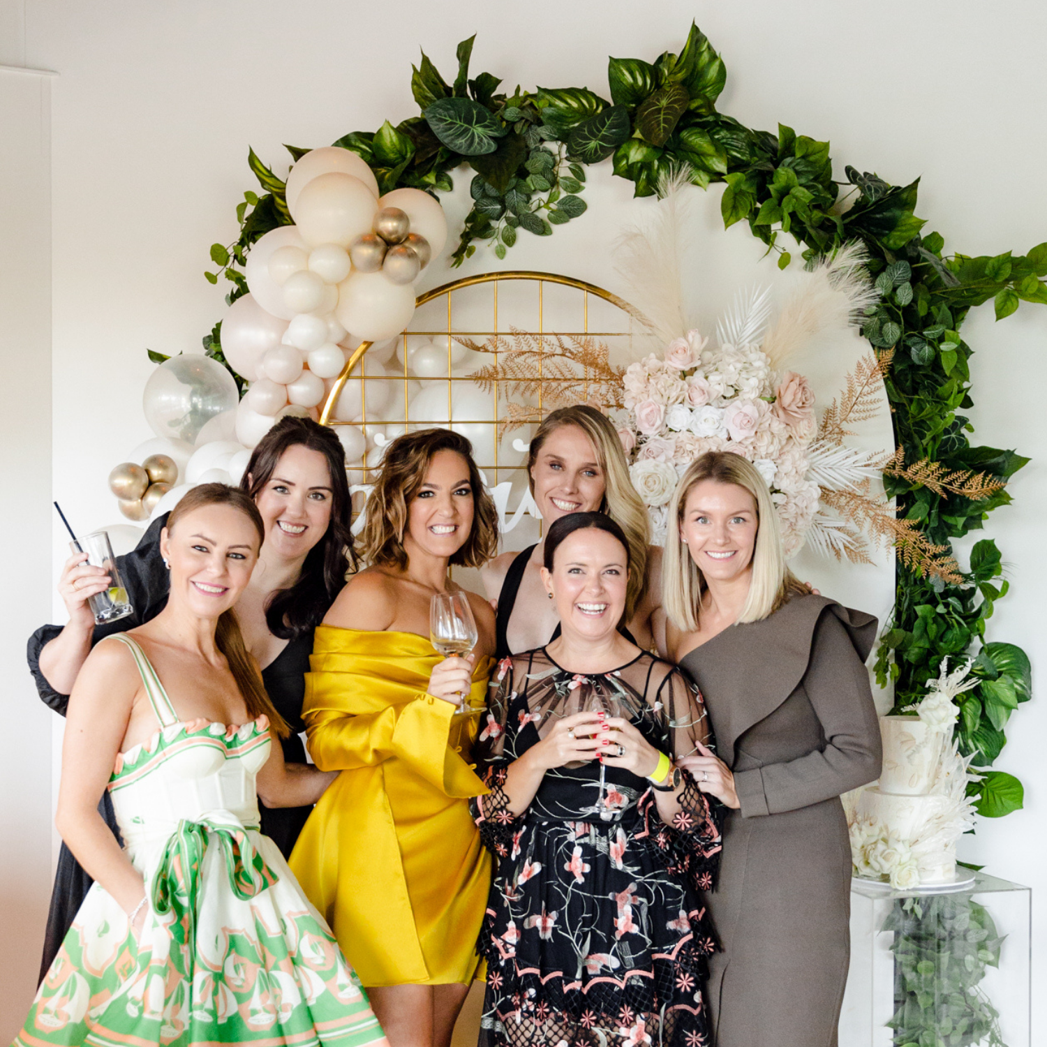 Group of seven women in colorful dresses and stylish outfits celebrating at a special event with floral and balloon decorations.