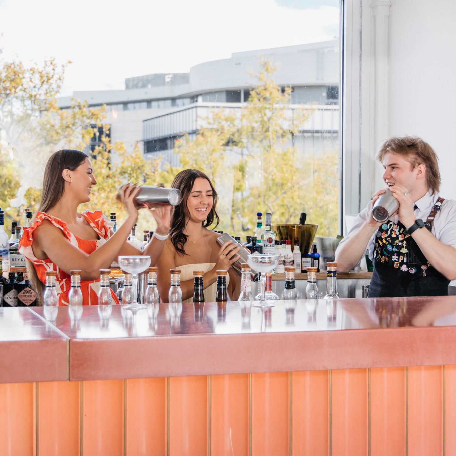 Three bartenders preparing cocktails at a bar, with bartenders smiling and mixing drinks, bottles of liquor and bar tools on the counter, large windows showing outdoor trees and buildings in the background.