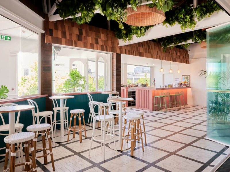 Modern cafe interior with white and wooden high tables and stools, large windows, green plants, a pink bar counter with green bar stools, and decorative greenery on the ceiling.