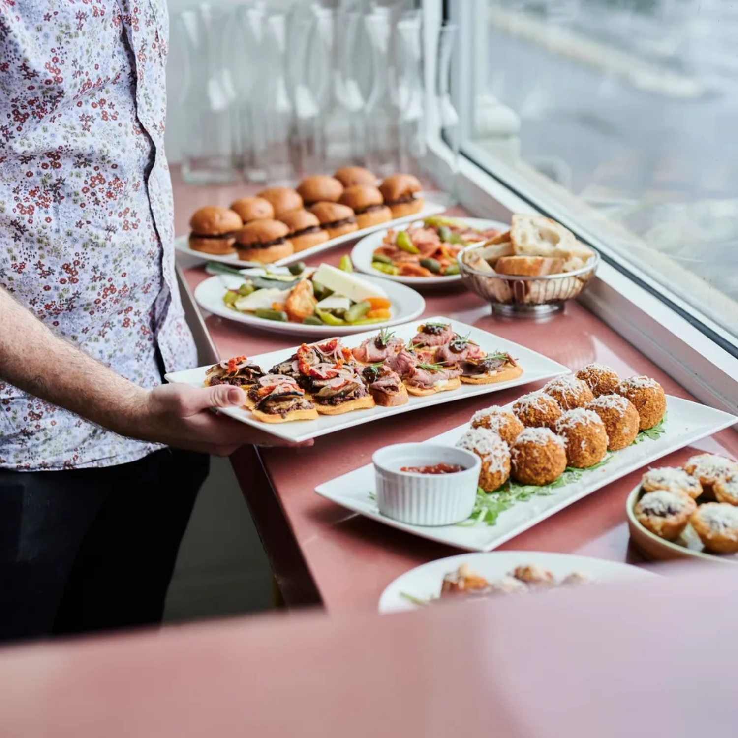 Person holding a plate of assorted appetizers and snacks at a buffet table with various finger foods, sandwiches, and desserts.