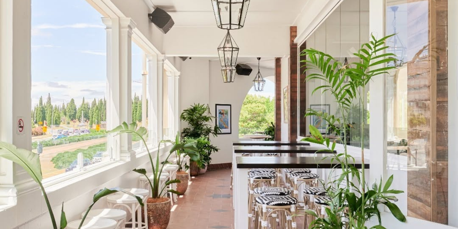 Bright indoor cafe or restaurant with large windows, leafy green plants in pots, bar-height tables with patterned stools, framed artwork on the wall, and pendant light fixtures, overlooking a green parking lot and landscape outside.