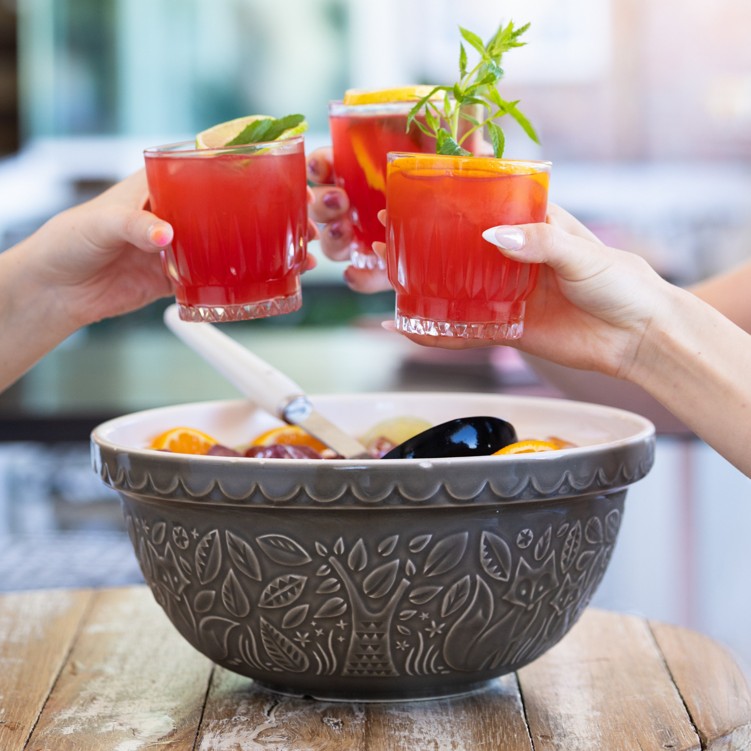 Three people raising glasses of red fruit punch with lemon slices and mint leaves for a toast over a large bowl of fruit salad.