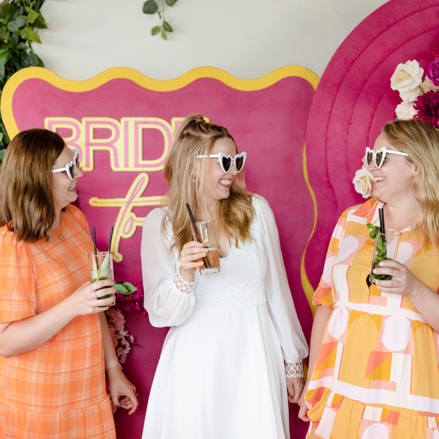 Three women at a bridal shower, wearing heart-shaped sunglasses, holding drinks, laughing and talking in front of a pink backdrop with the words 'Bridal' visible.
