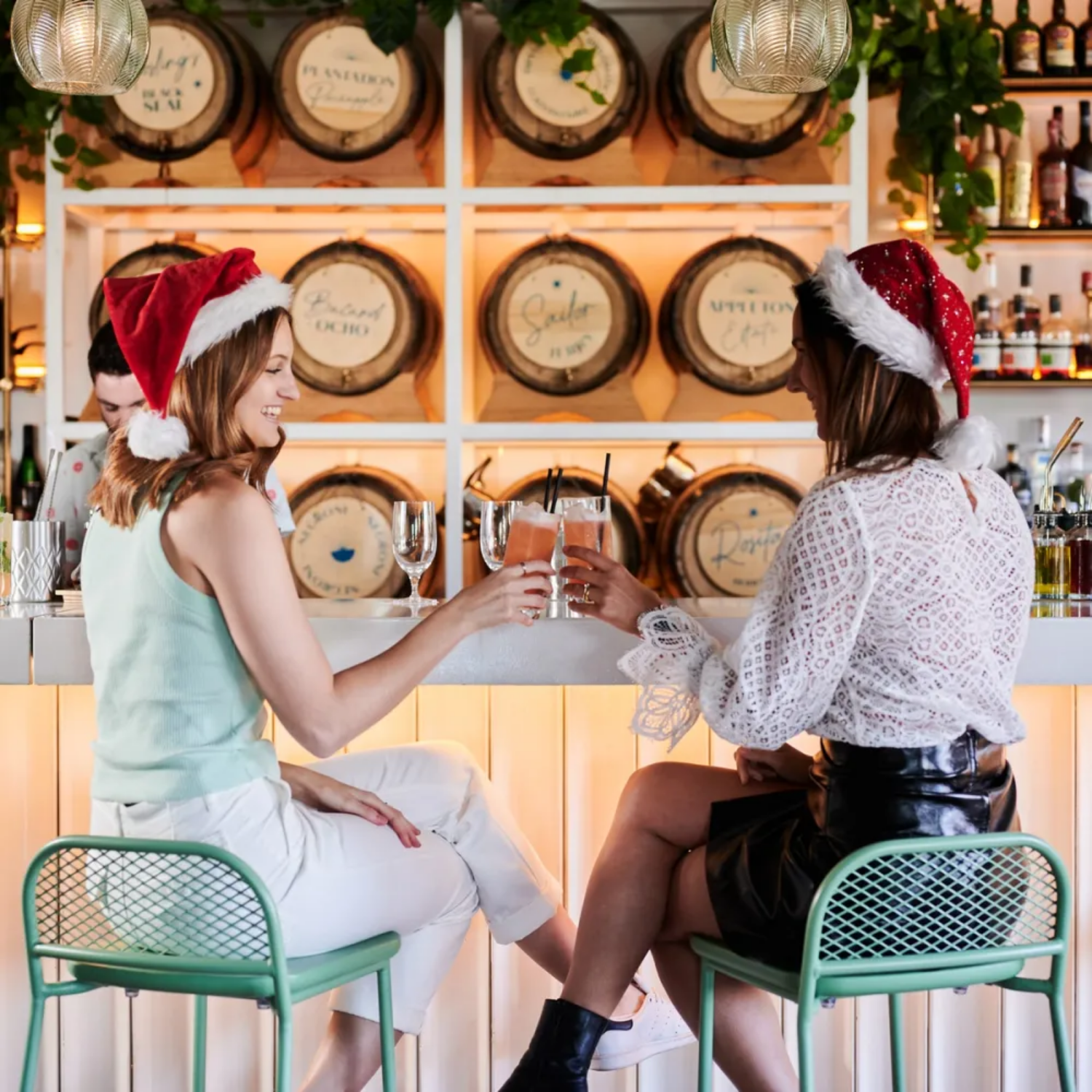 Two women sitting at a bar wearing Santa hats, clinking glasses with cocktails, celebrating Christmas.