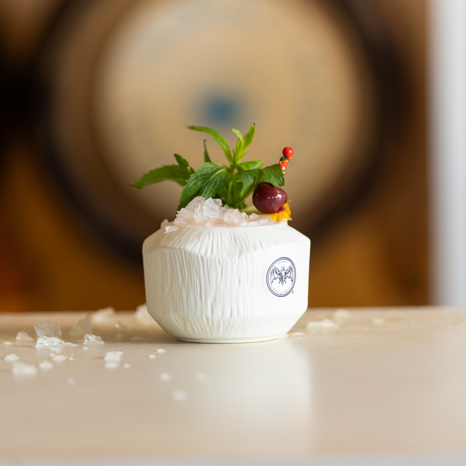 A white ceramic dish with a logo, topped with ice, garnished with a sprig of green mint and a cherry, with small scattered ice pieces on a white surface, blurred wooden object in the background.