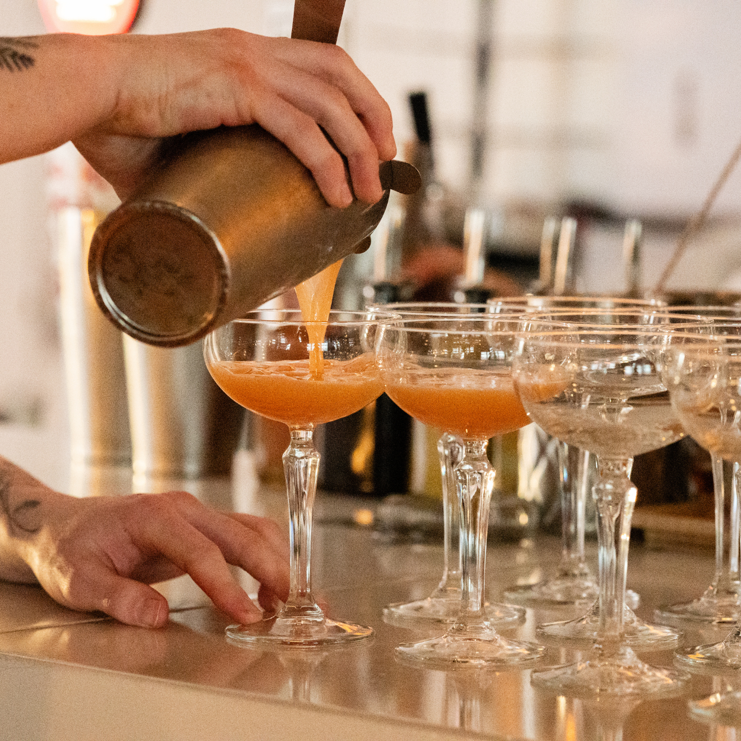 A bartender is pouring a pink cocktail from a shaker into glasses on a bar.