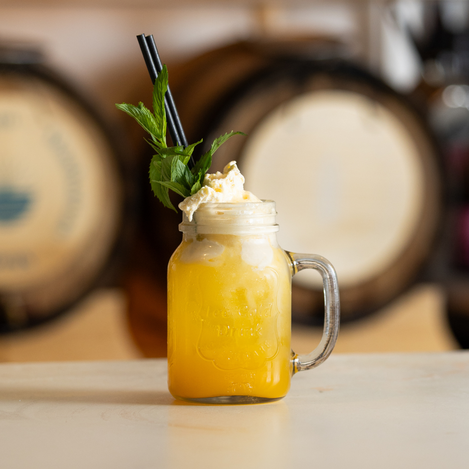 A yellow beverage topped with whipped cream, garnished with mint leaves, with two black straws in a glass mason jar, set against a blurred background of bourbon barrels.