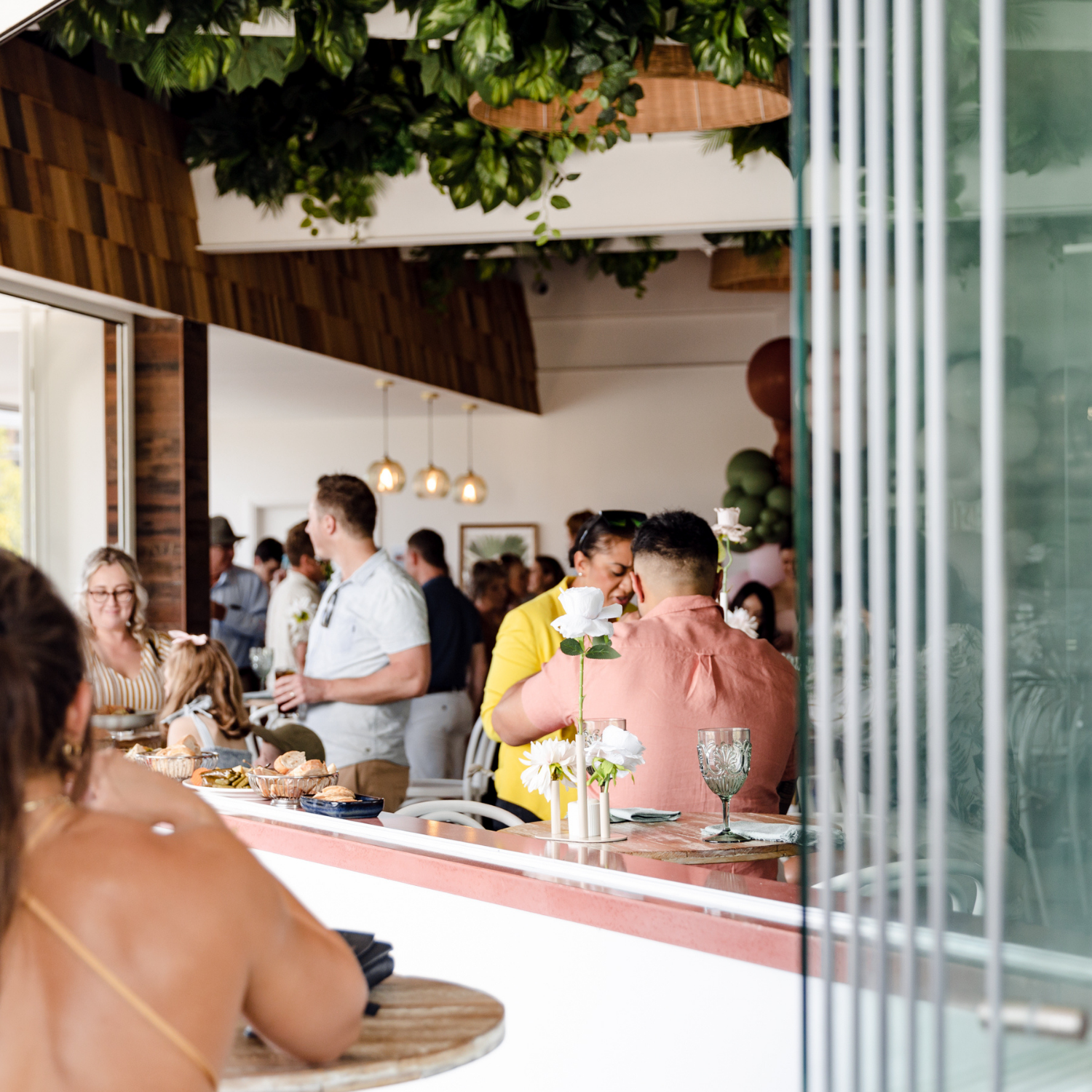 People socializing at a lively indoor gathering, with a bar area, floral decor, and a modern, airy interior.