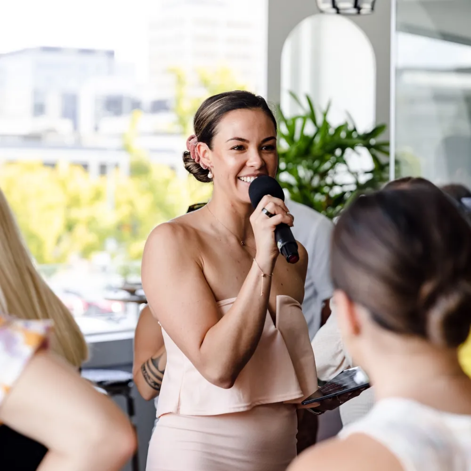 A woman in a strapless beige dress smiling and speaking into a microphone at an indoor event with large windows and greenery outside.