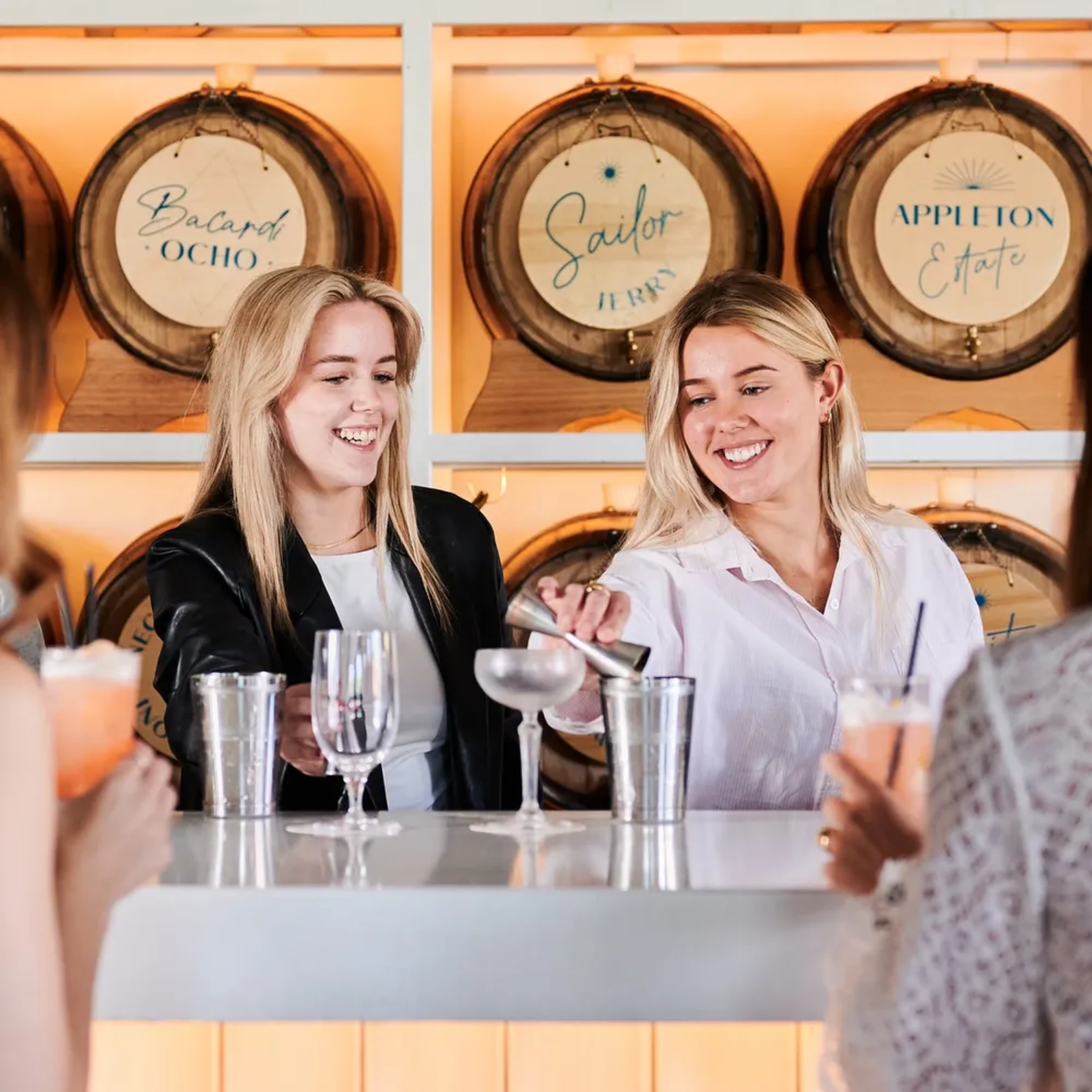 Two women behind a bar counter smiling and having fun, with champagne glasses and cocktails in front of them. They are in a setting with wooden barrels in the background that have signs reading 'Bacard Ocho,' 'Sailor Jerry,' and 'Appleton Estate.'