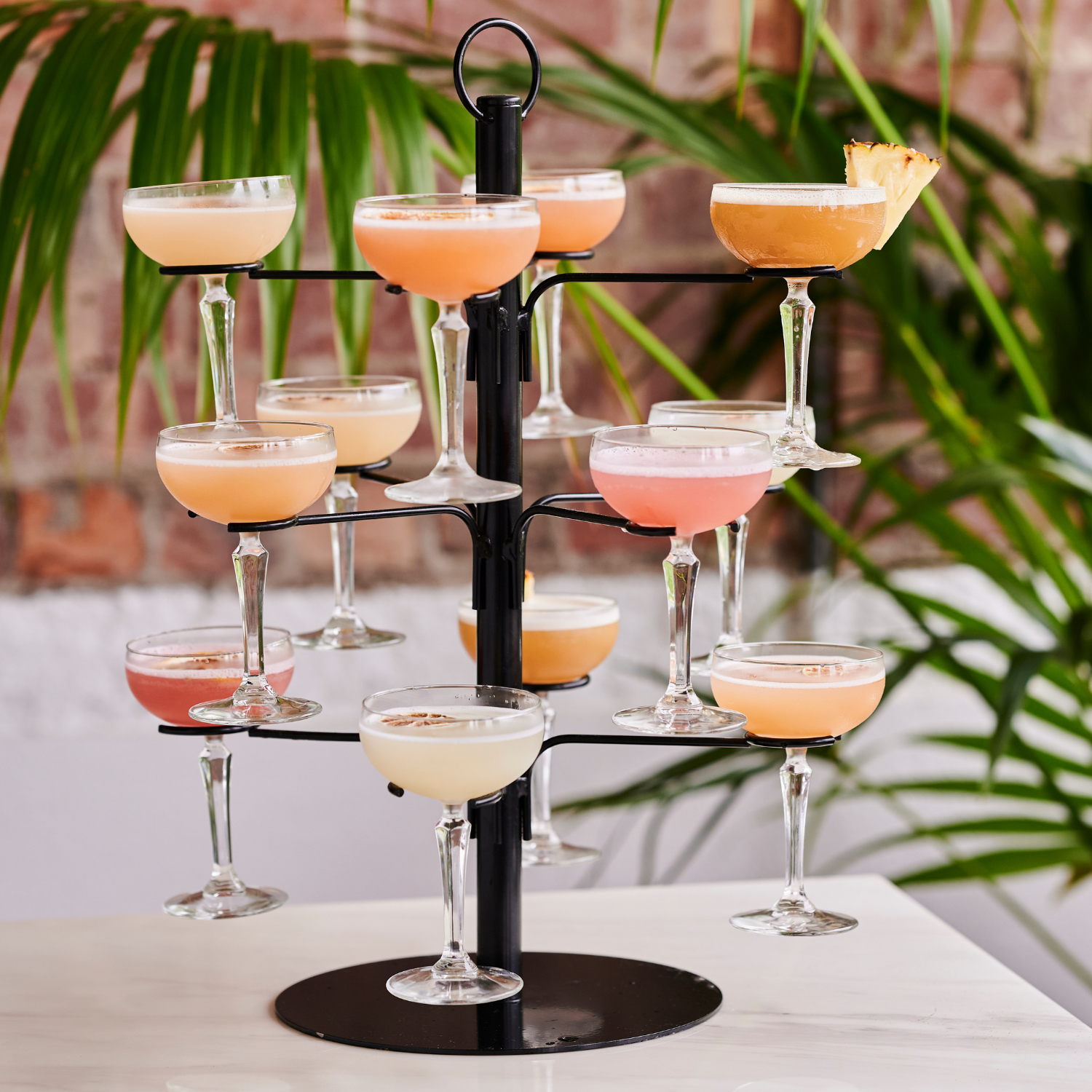 A tiered black stand holding multiple cocktail glasses filled with colorful drinks in pink, cream, and orange hues, some garnished with fruit, set against a background of green plants and a brick wall.