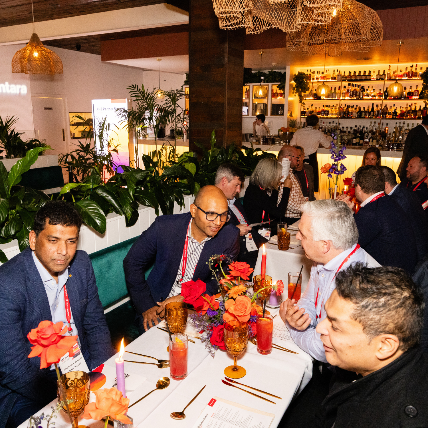 People sitting at a decorated dining table with flowers and drinks, engaging in conversation at a social event in a bar or restaurant with a bar counter in the background.