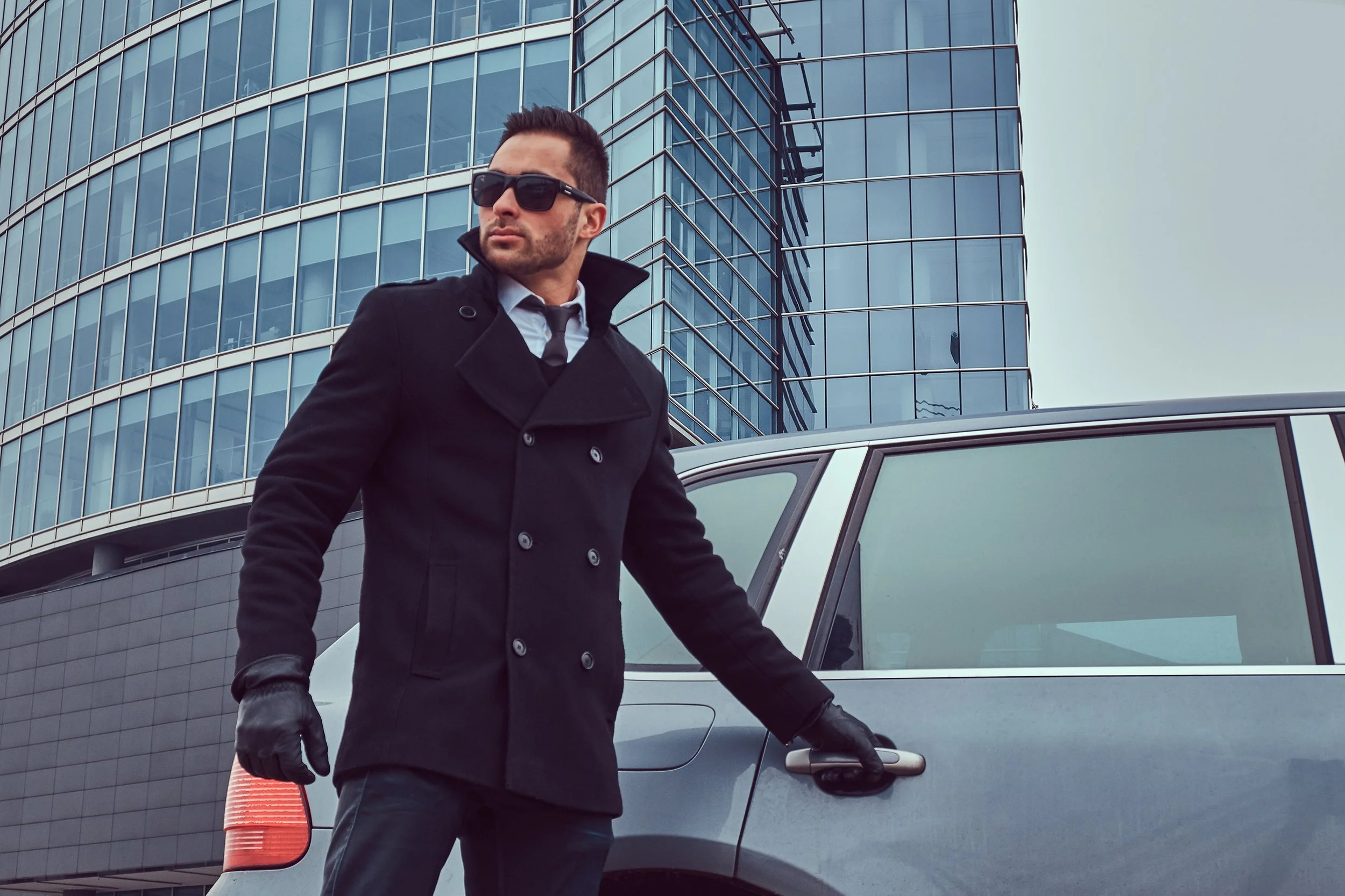 A man wearing a black coat, sunglasses, and gloves stands next to a silver SUV with a modern office building in the background.