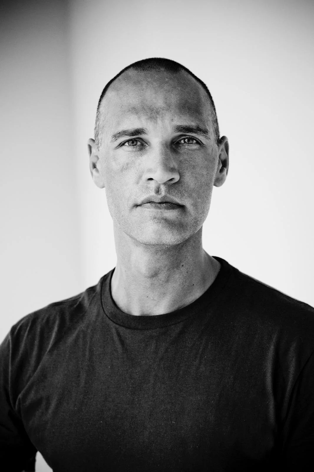 Black and white portrait of a man with short hair wearing a black T-shirt, looking directly at the camera.