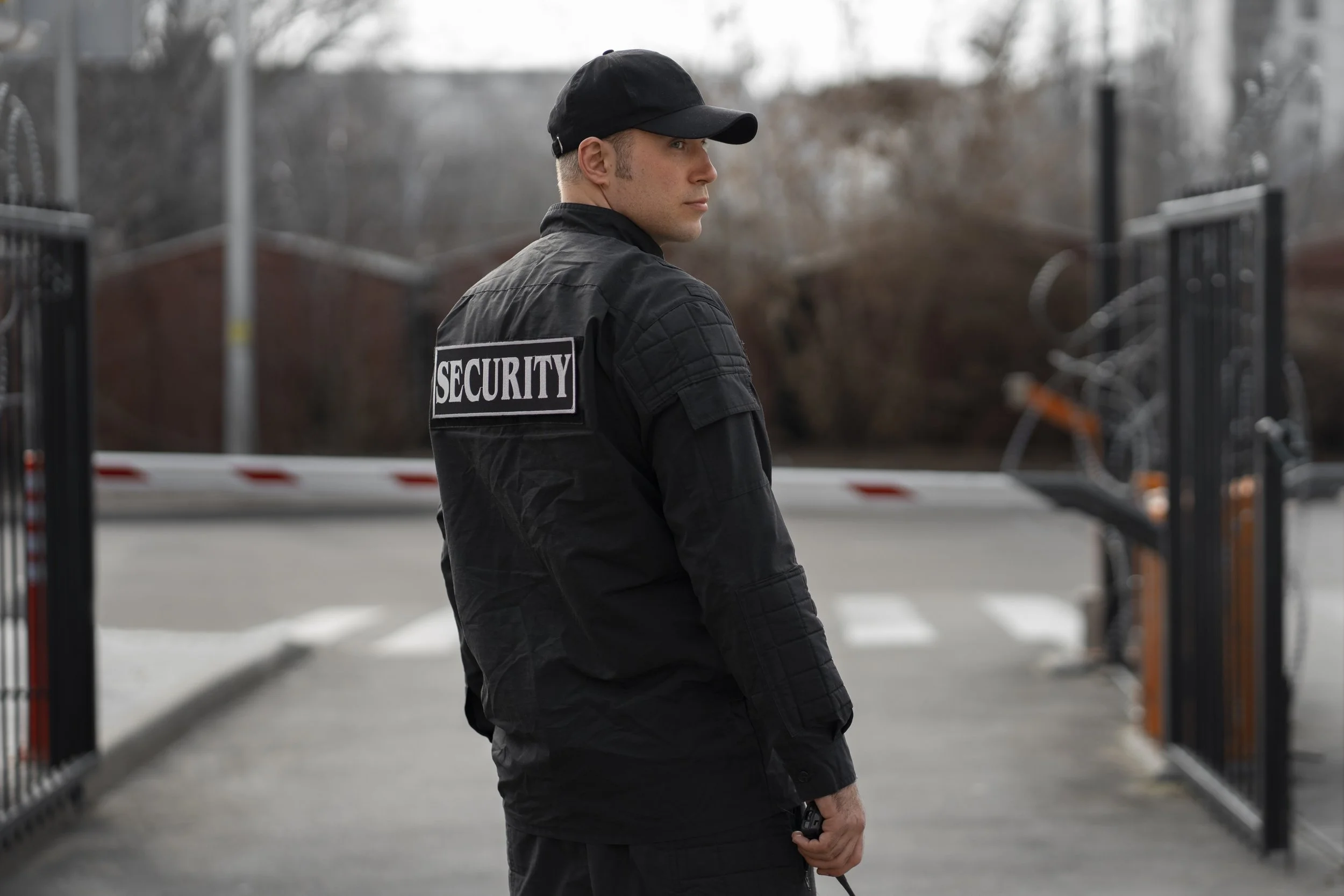 A security guard stands outdoors near a closed gate, holding a walkie-talkie, wearing a black uniform with a "SECURITY" patch on the back, and a black cap.