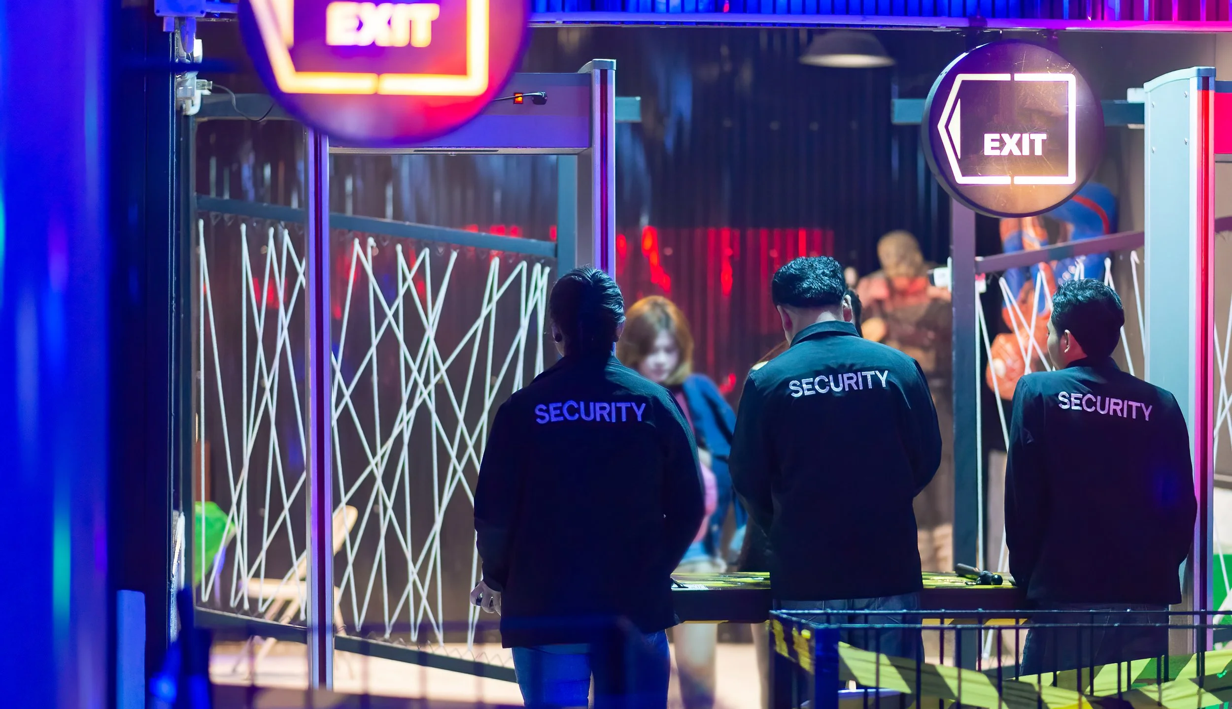 Three security guards stand at an entrance with neon exit signs and a crowd in the background at a dimly lit venue.