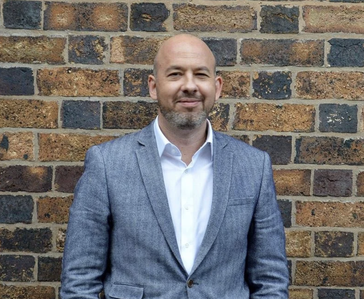 A man with a beard wearing a gray blazer and white shirt standing against a brick wall.