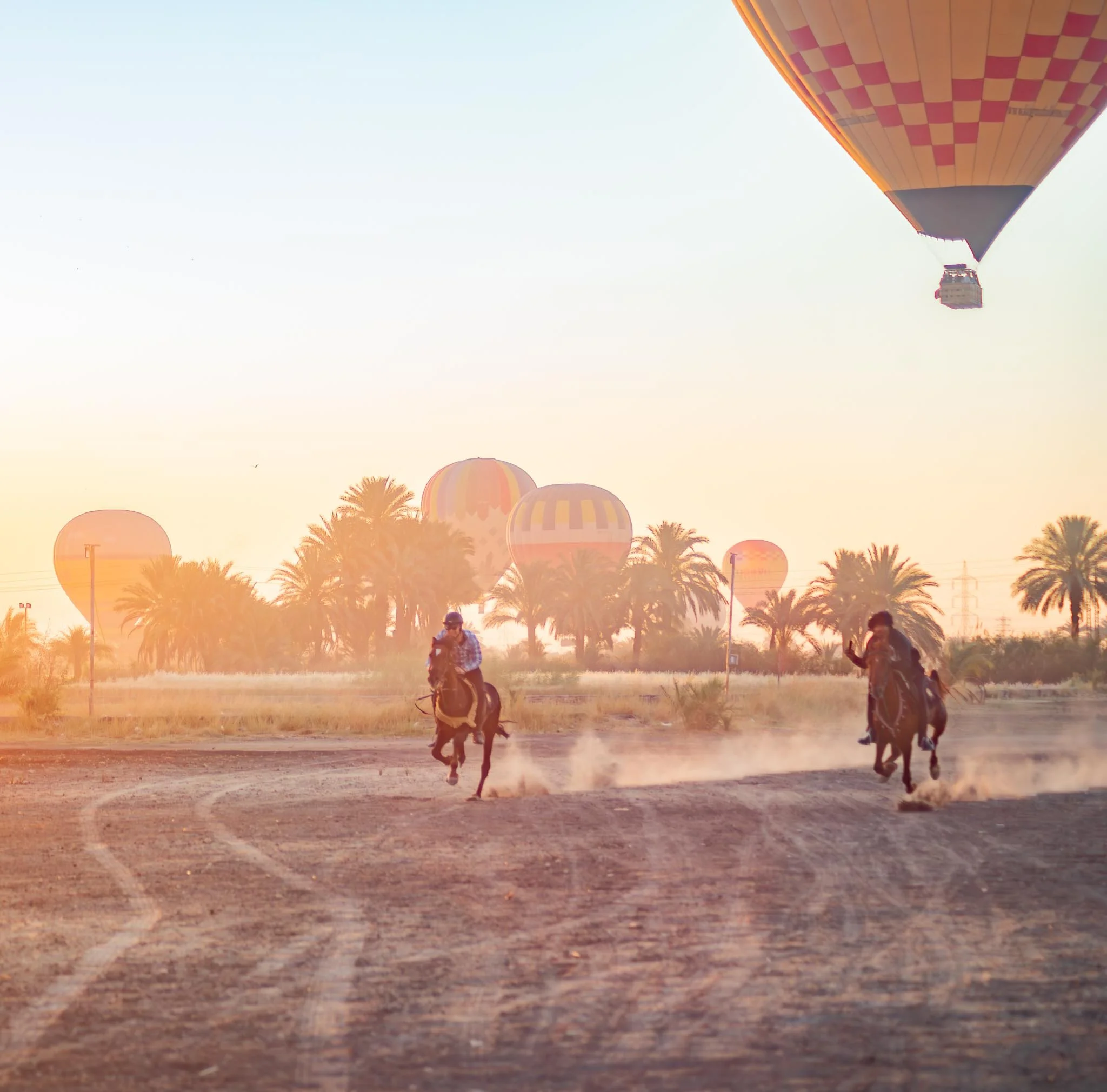 A hot air balloon with orange and pink checkered pattern flying in the sky during sunset, with palm trees and people on the ground.