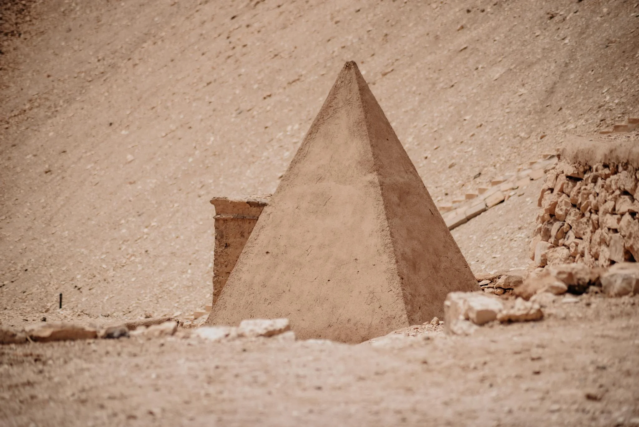A small, sandy pyramid-shaped structure on a desert-like surface, at the tomb of the scribes in Luxot, with a flat base and a pointed top, surrounded by scattered rocks and gravel.