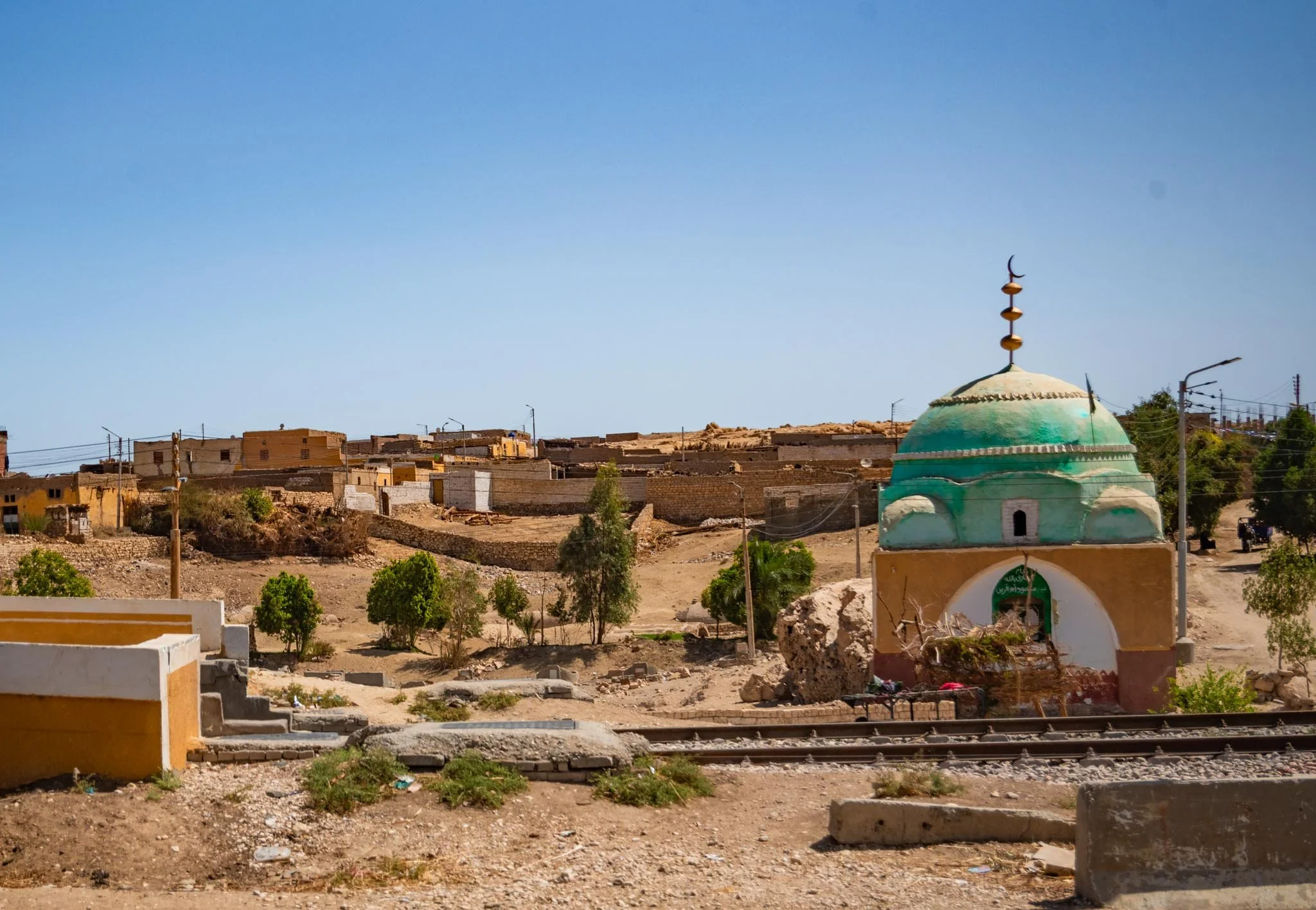 A colorful mosque with a turquoise dome and a crescent moon finial, set against a clear blue sky in a small town with low-rise buildings and greenery on the road to Aswan