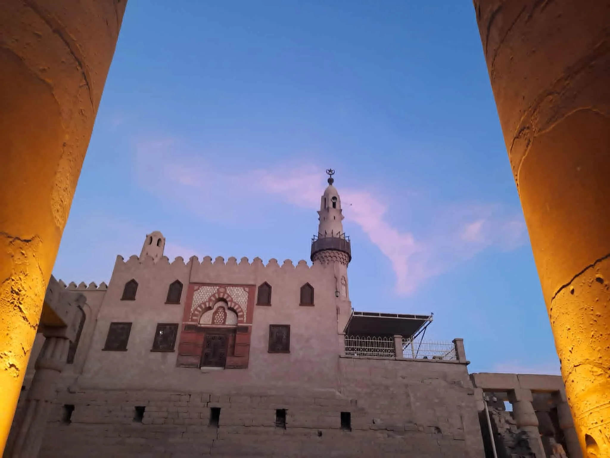 view of the mosque in the Luxor Temple seen through a frame of two large, yellow, textured pillars at dusk. The building has decorative arches and a tall minaret topped with a crescent moon symbol, set against a blue evening sky.